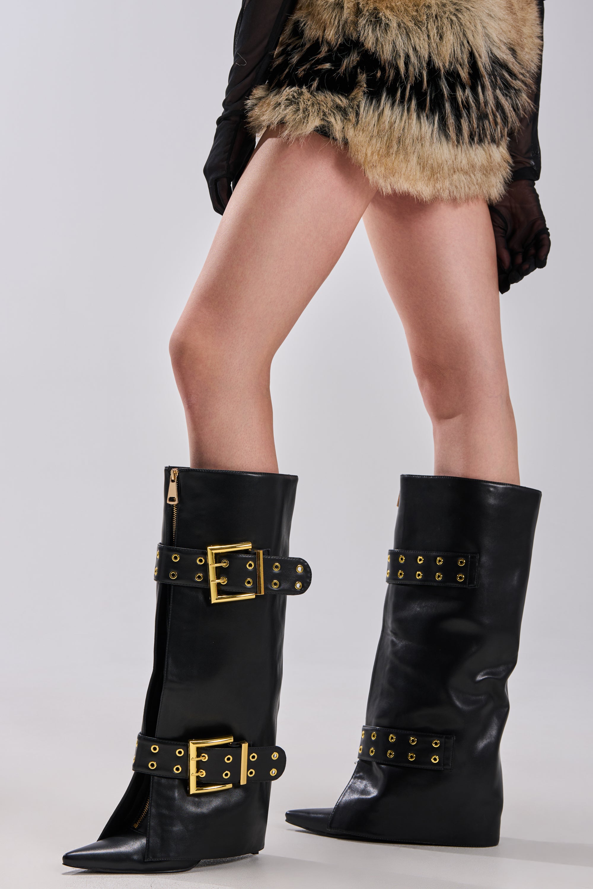 A person wears the AZALEA WANG ROOVER BLACK HARDWARE EMBELLISHED STILETTO BOOT, featuring large gold buckles and grommet details, styled with a fur-textured skirt and black mesh gloves.