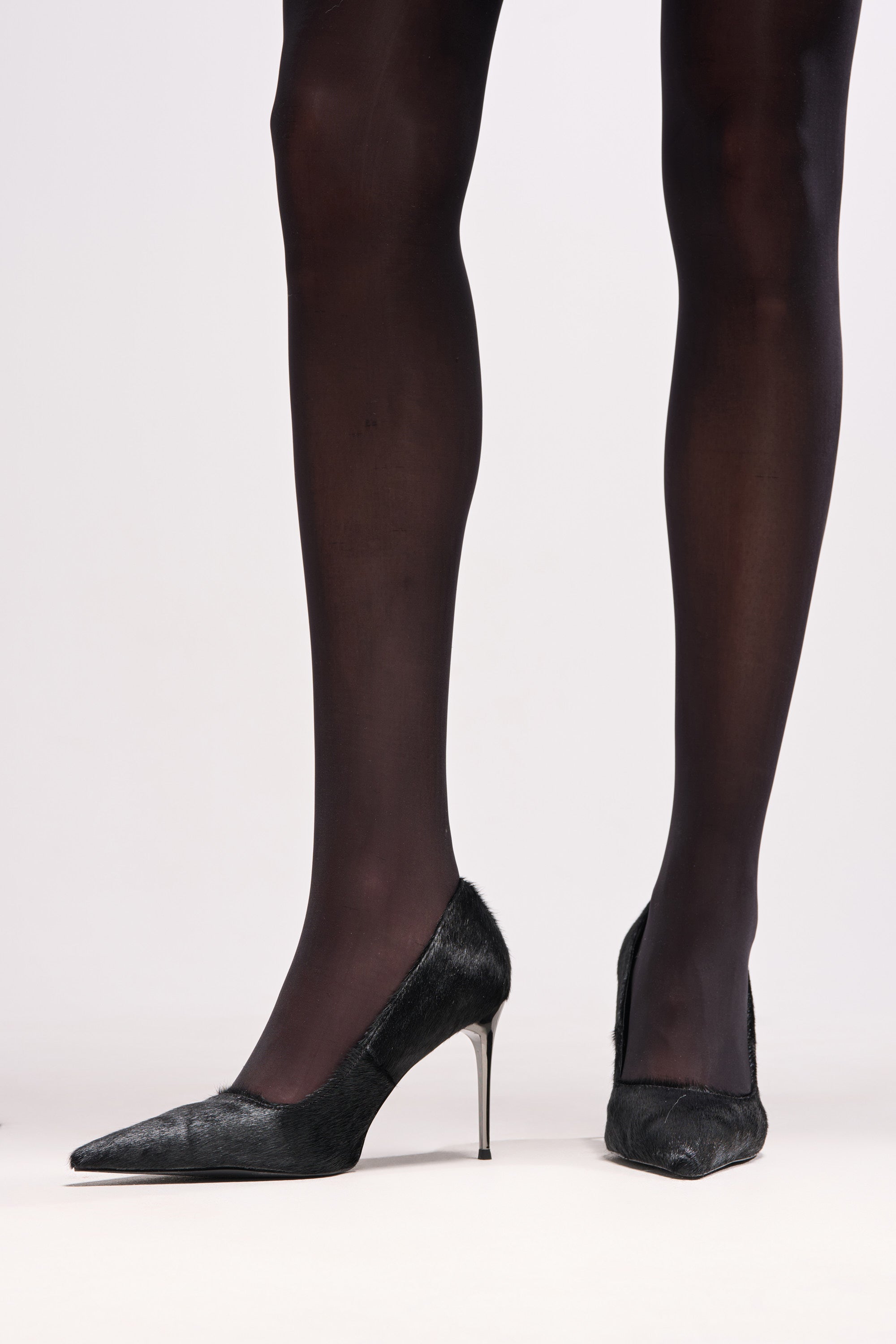 A person wearing sheer black tights and the AZALEA WANG ROSEE BLACK PONY HAIR PUMP with pointed toes and stiletto heels stands against a light background.