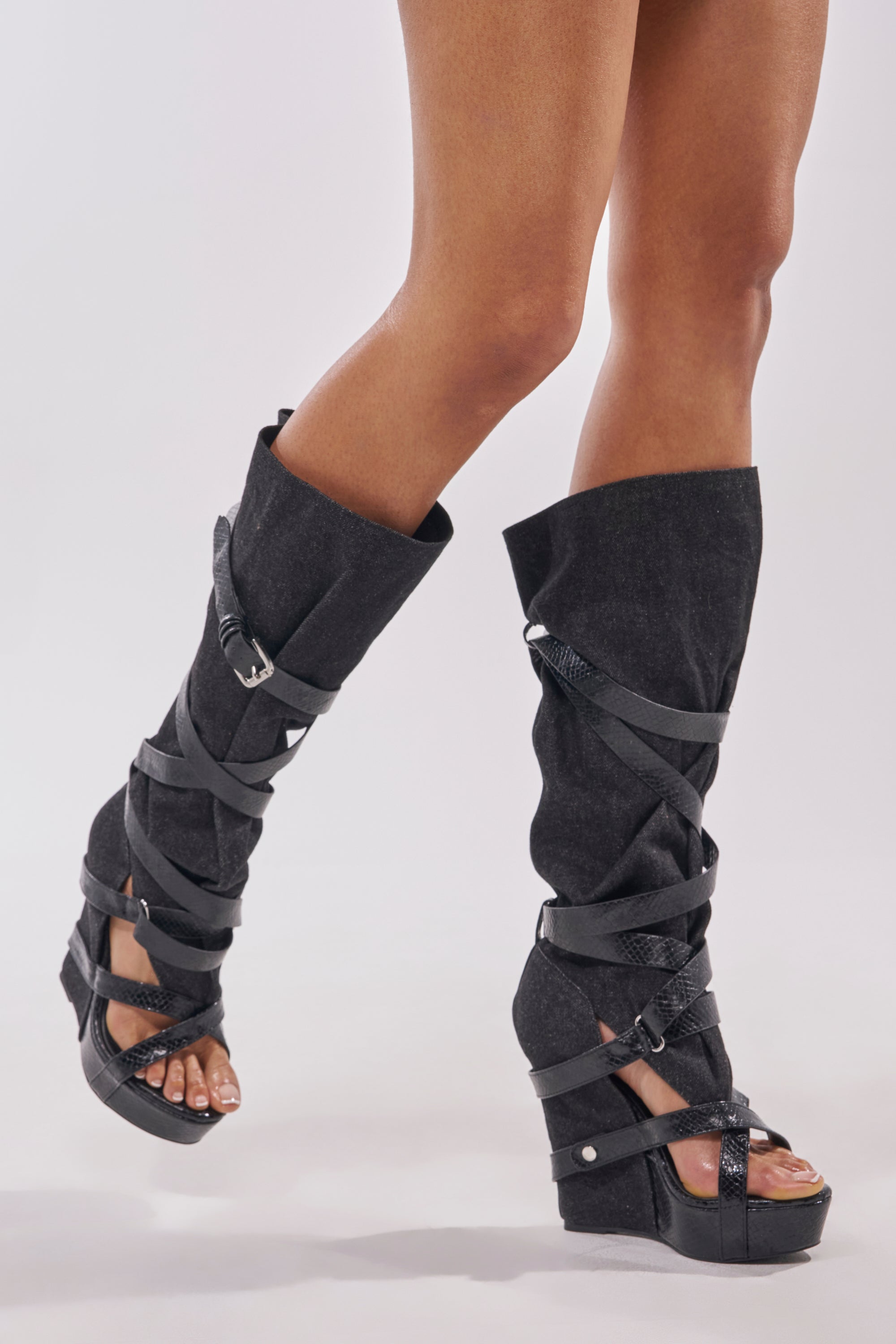 A person wearing the AZALEA WANG ROTHKID BLACK DENIM KNEE HIGH OPEN TOE BOOT stands with one leg crossed in front of the other against a plain background, showing off the black denim open-toe and exposed heel style. 7
