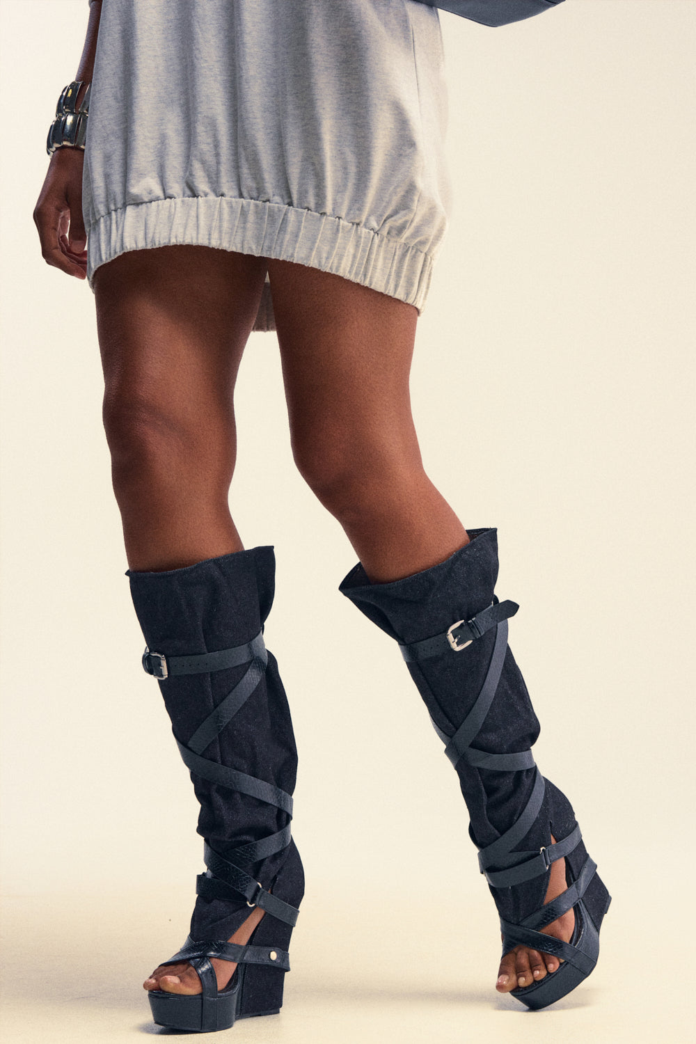 A person with dark skin wears a short, light-gray gathered-hem dress paired with statement AZALEA WANG ROTHKID Black Denim Knee High Open Toe Boots. The boots feature dark denim fabric, wraparound straps, buckles, and an open-toe silhouette. Isolated on white.