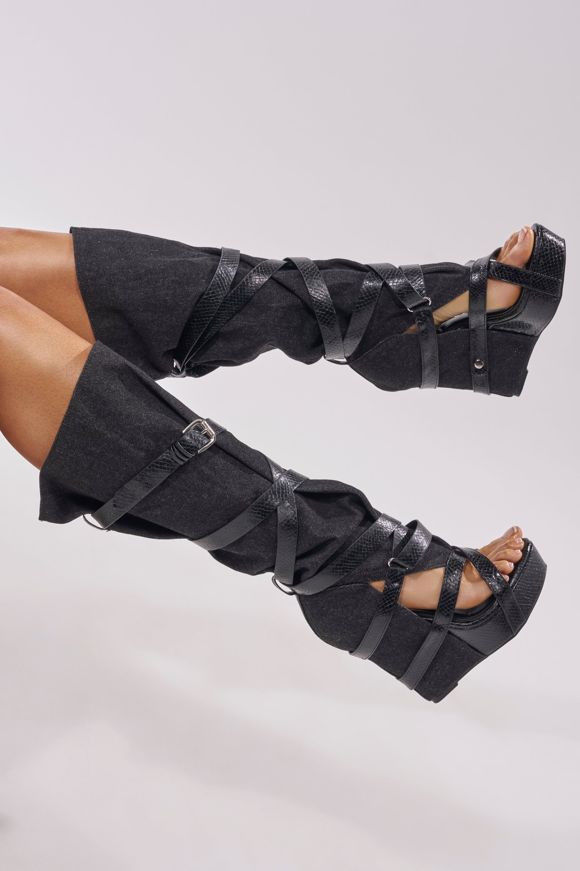 A pair of legs stylishly raised in AZALEA WANG ROTHKID BLACK DENIM KNEE HIGH OPEN TOE BOOTS, featuring black wedge heels and crisscrossing black straps, set against a plain light background. 3