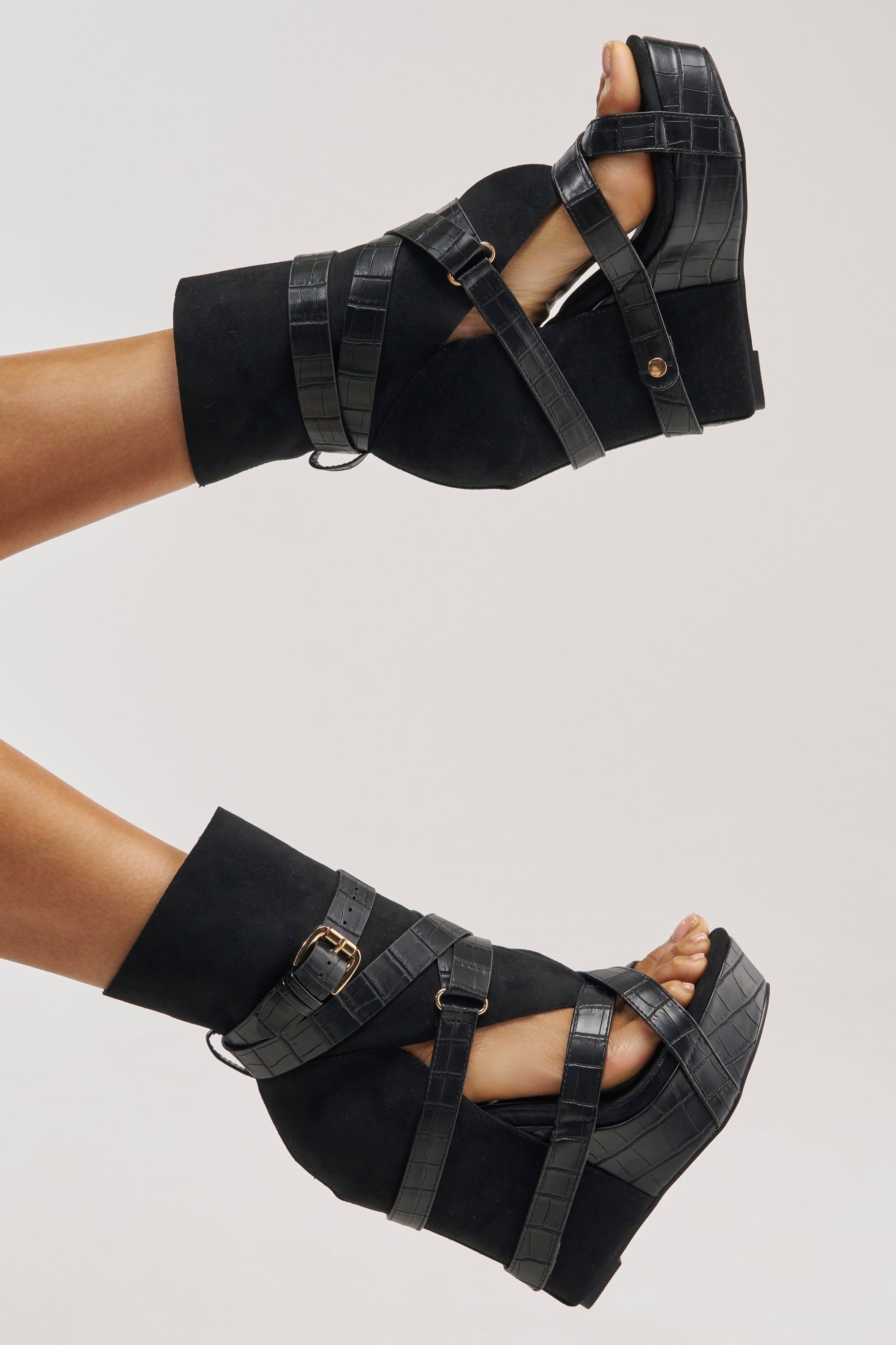 A person's legs are shown raised, wearing the AZALEA WANG ROTHKO WEDGE SANDAL IN BLACK. These sandals showcase a bold platform sole, faux crocodile-textured straps reminiscent of a black gladiator style, a suede-like base, and gold-toned buckles, all set against a plain light gray background.