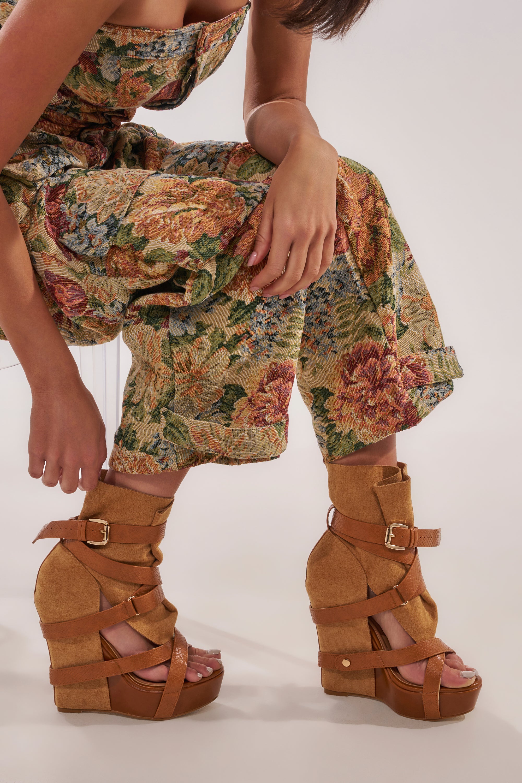 A woman in a floral jumpsuit wears AZALEA WANG ROTHKO WEDGE SANDALS IN CAMEL, featuring crisscross straps and buckles, as she sits with bent knees and one hand on her leg against a plain background. 3
