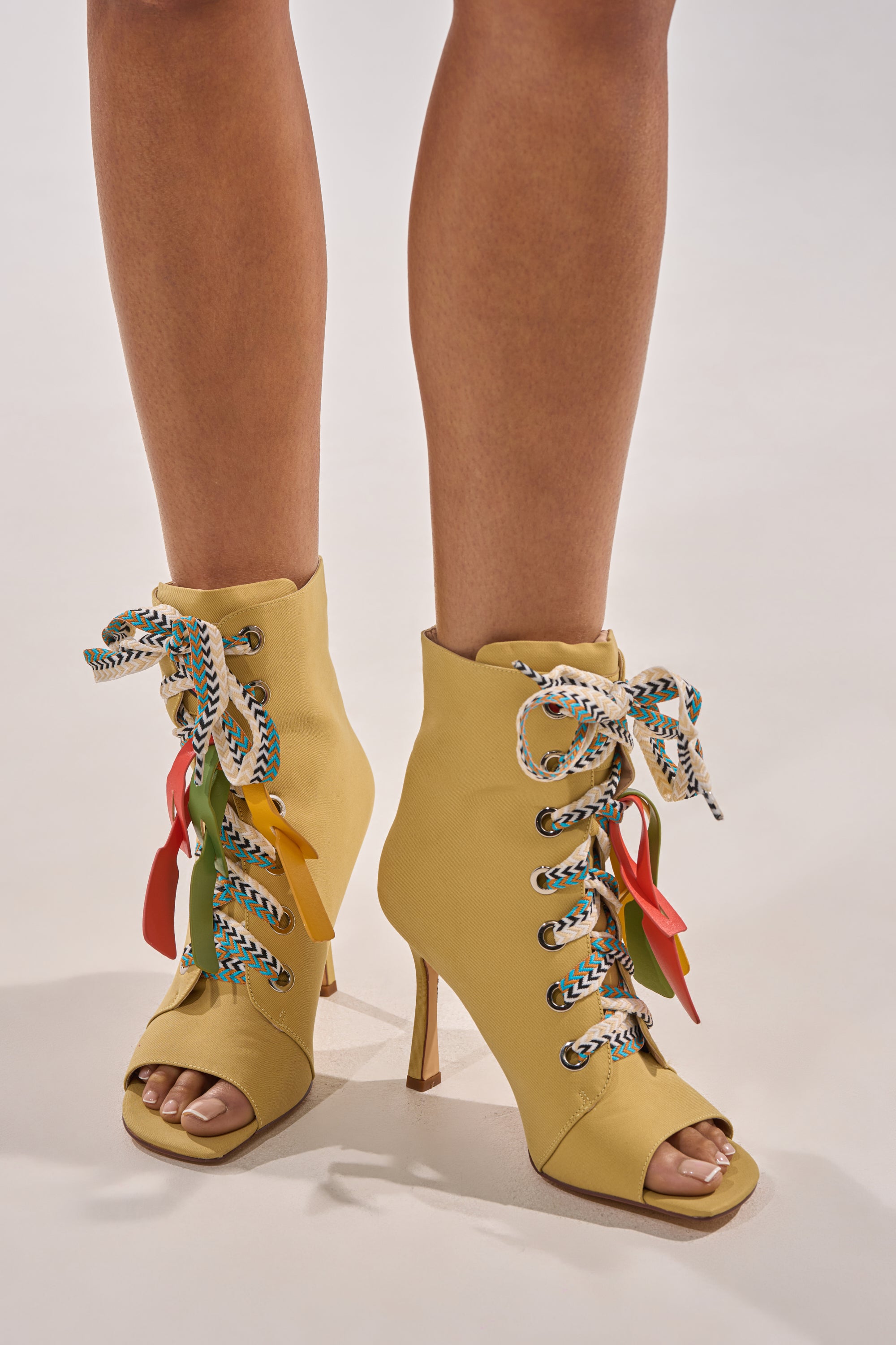 A person stands on a light background wearing the AZALEA WANG ROVEA KHAKI LACE UP STILETTO BOOTIE, featuring yellow color, open toes, multicolored patterned laces, and decorative leaf-shaped charms. 2