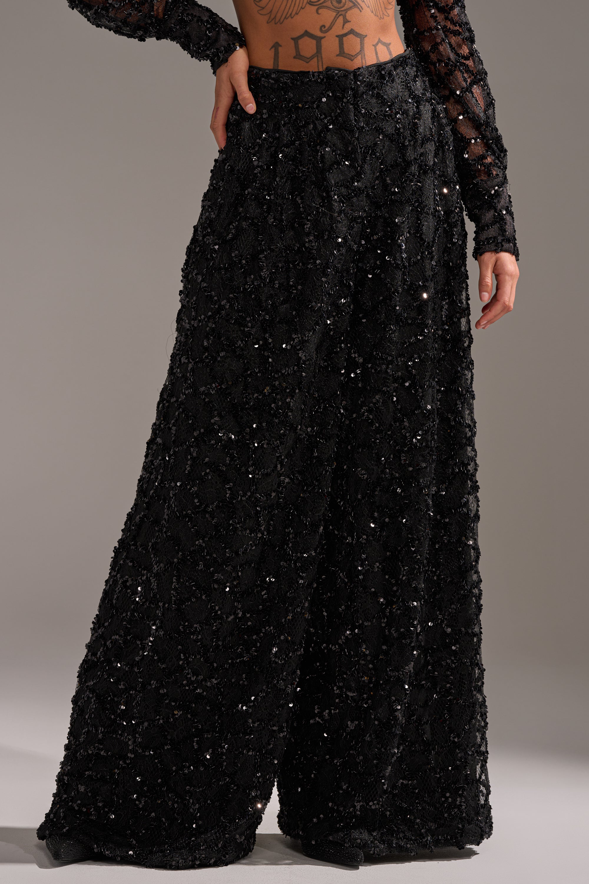 A person stands against a plain background wearing ROXY SEQUIN PALAZZO PANT, featuring a high waist and wide-leg silhouette, paired with a mesh long-sleeve top that reveals midriff tattoos.