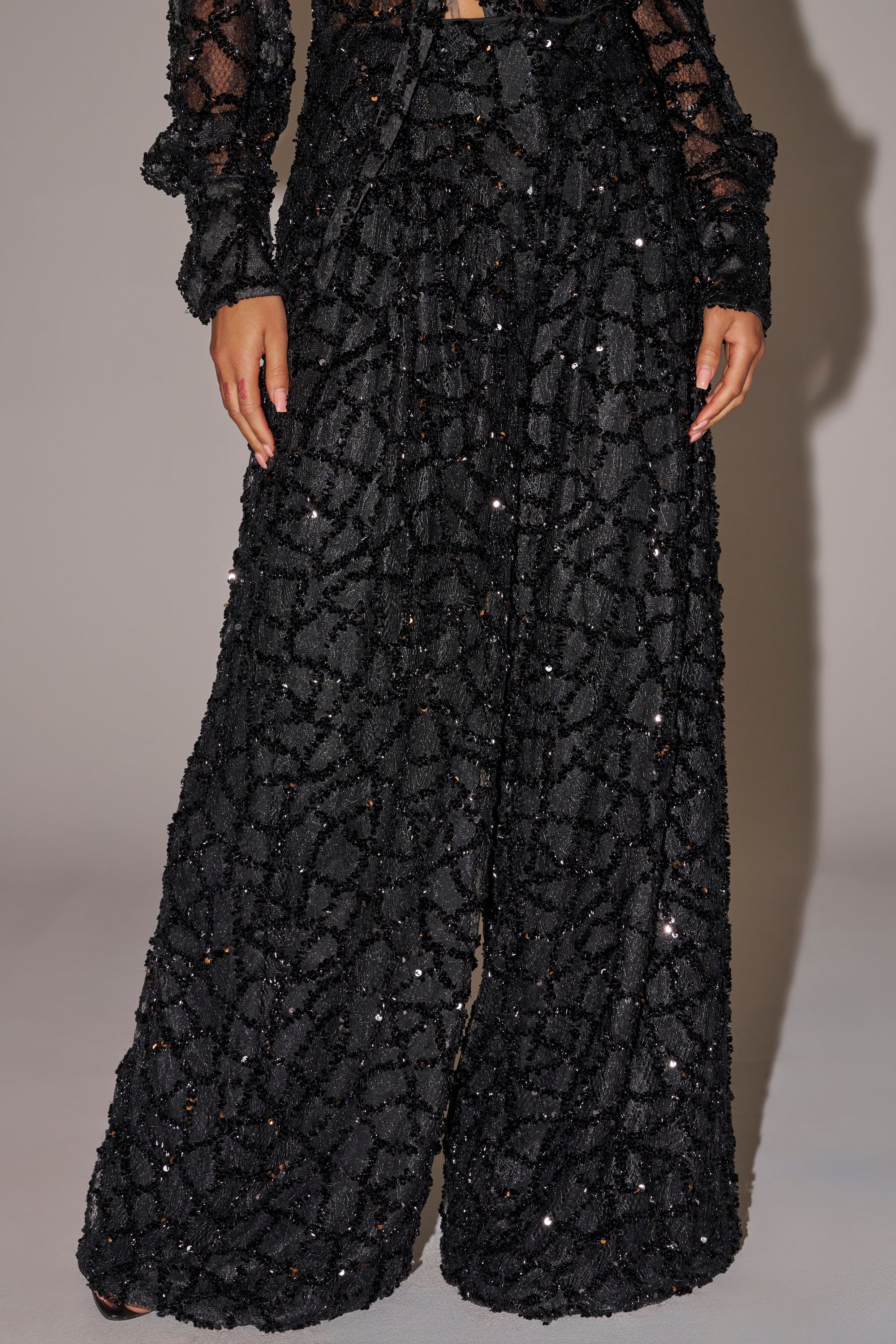 Wearing the ROXY SEQUIN PALAZZO PANT, a high waisted, textured, glittering wide-leg silhouette paired with a matching long-sleeved top; only the body from mid-torso down is visible. 1