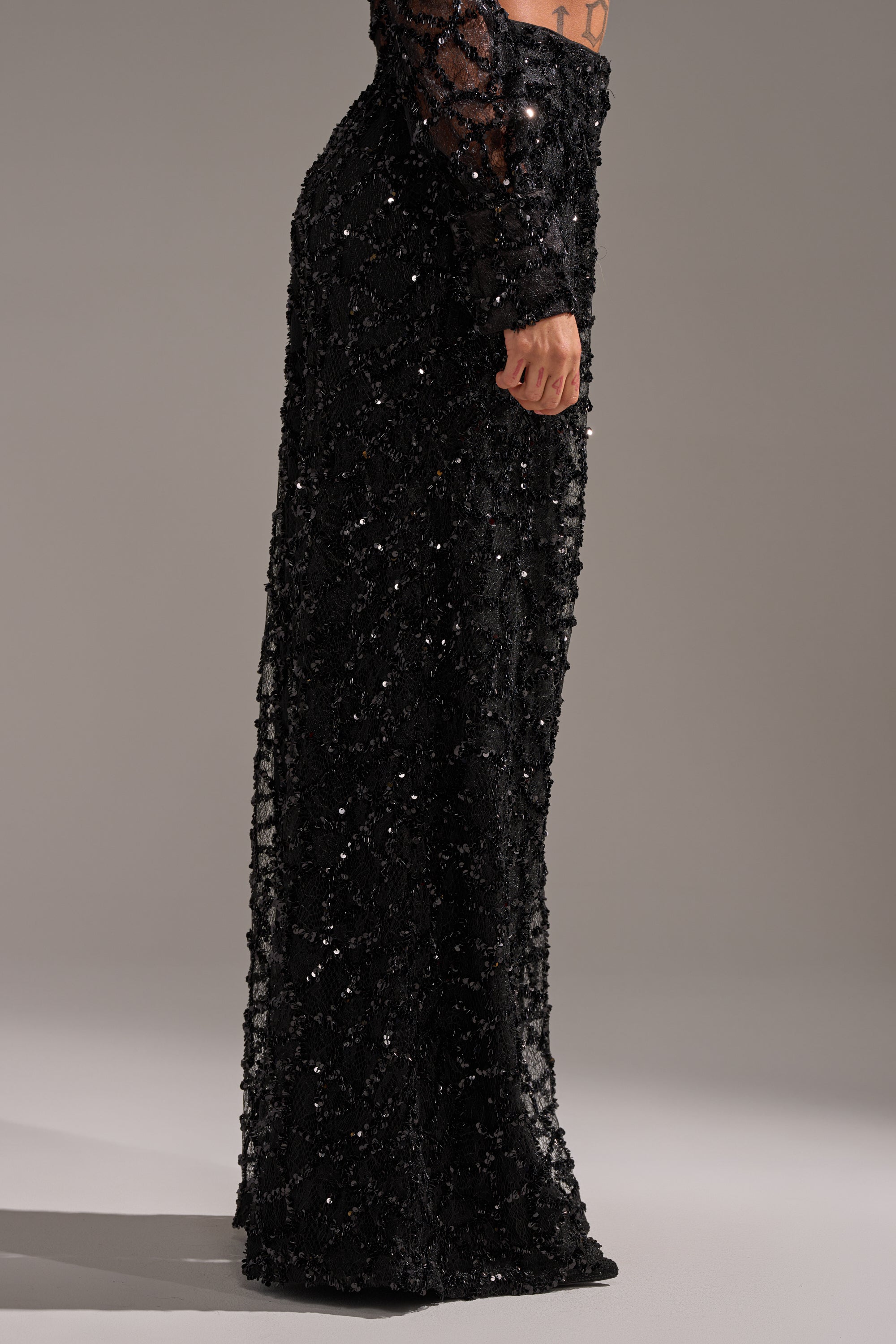 A person stands sideways in the ROXY SEQUIN PALAZZO PANT, featuring a fitted black sequin design with a sheer, sparkling overlay and wide-leg silhouette. Their arm is relaxed by their side against a plain, neutral background.