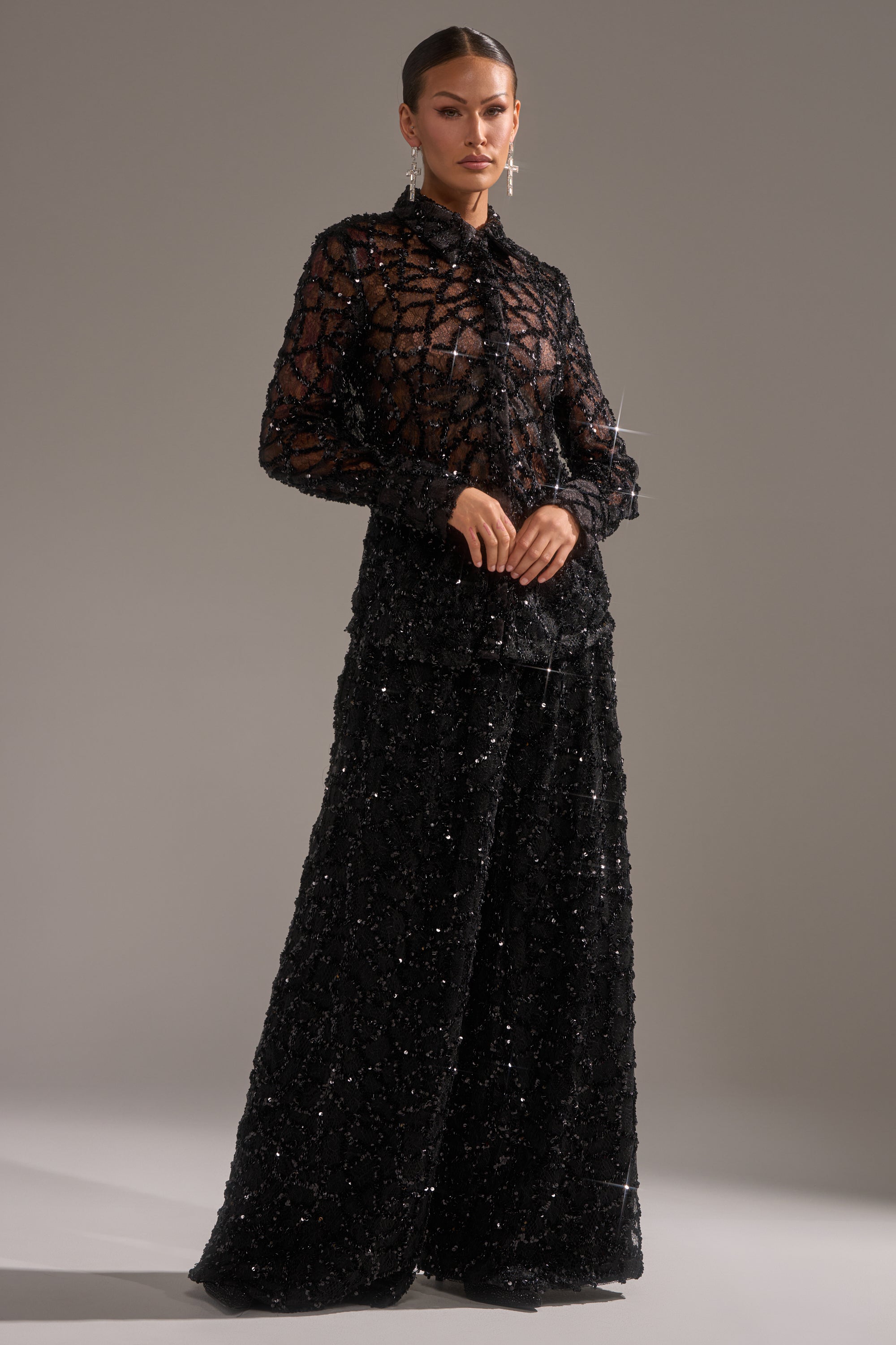 A woman poses against a plain background, wearing the ROXY SEQUIN PALAZZO PANT—sparkling, wide-leg palazzo pants paired with a sheer black long-sleeve blouse. She accessorizes with earrings, slicked-back hair, and a neutral expression.