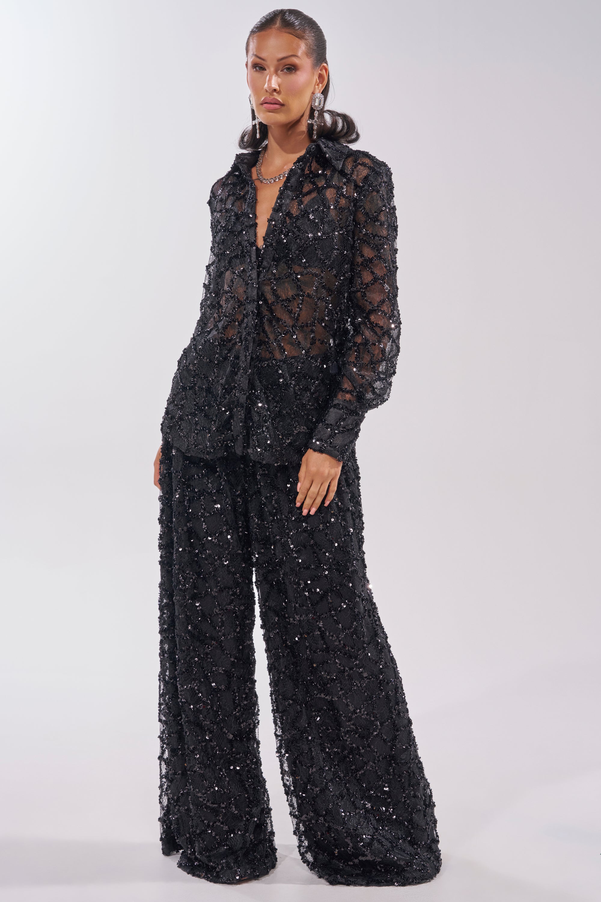 A woman poses against a plain light background, wearing the ROXY SEQUIN PALAZZO PANT—a black, sheer, sequin-embellished wide-leg pants set—styled with a matching blouse and gold jewelry. 7