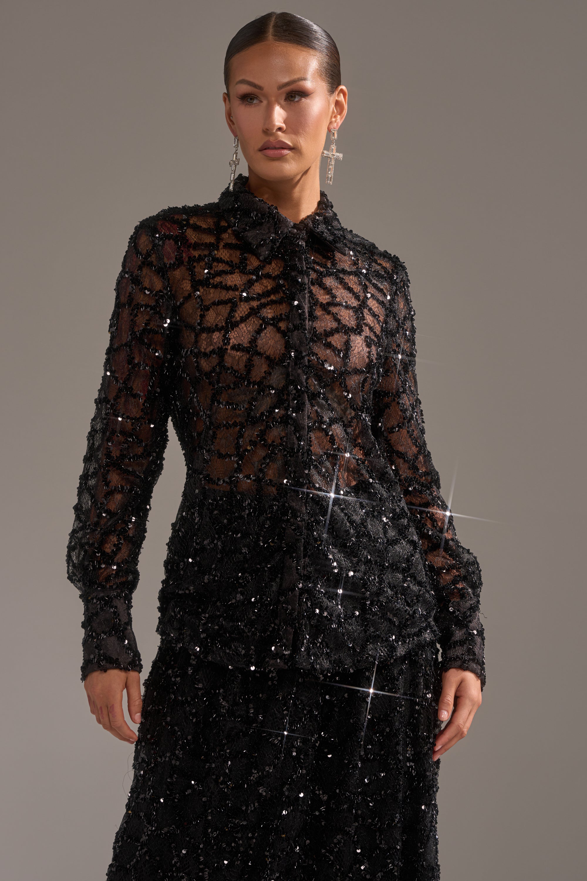 A woman wears the ROXY SEQUIN BUTTON DOWN blouse with a matching skirt. Her slicked-back hair and drop earrings finish the evening look as she stands against a plain background, expression neutral.