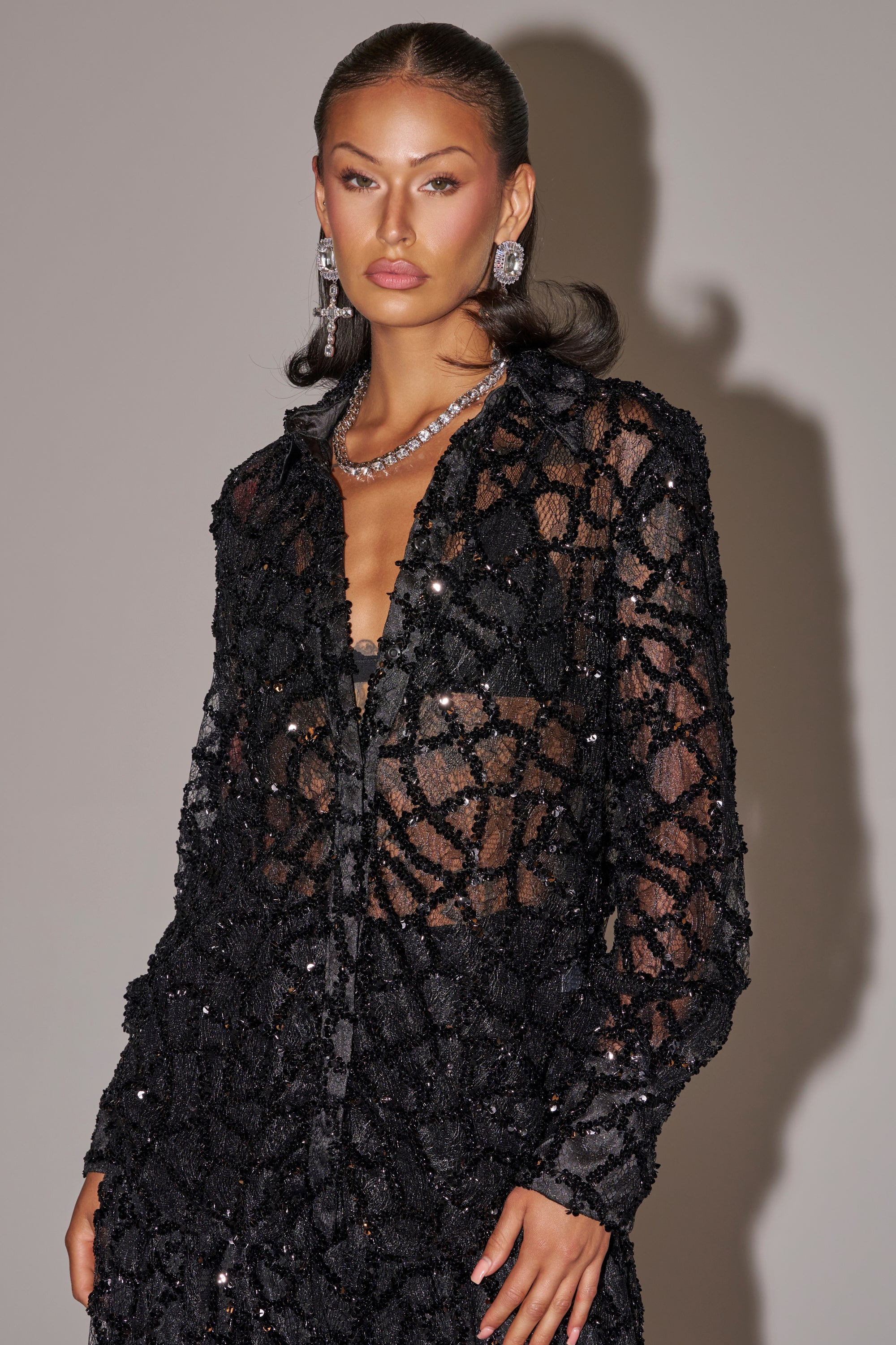 A woman with slicked-back dark hair wears the ROXY SEQUIN BUTTON DOWN over a black bra, paired with statement earrings and a silver necklace, creating a bold evening look against a plain light background.