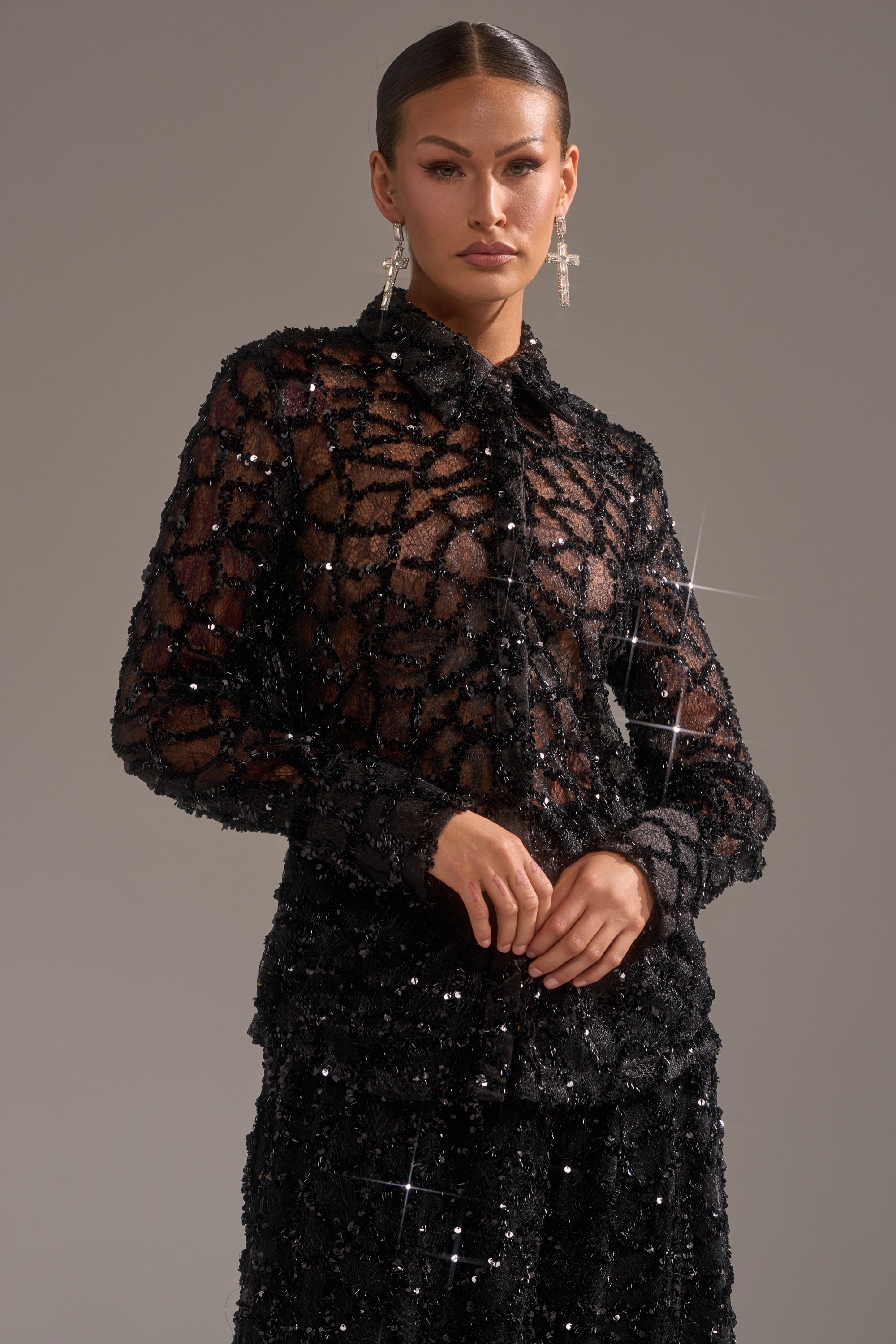 A woman with slicked-back hair models the ROXY SEQUIN BUTTON DOWN, paired with a matching skirt. She clasps her hands and wears bold earrings, creating an elegant evening look against a plain background.
