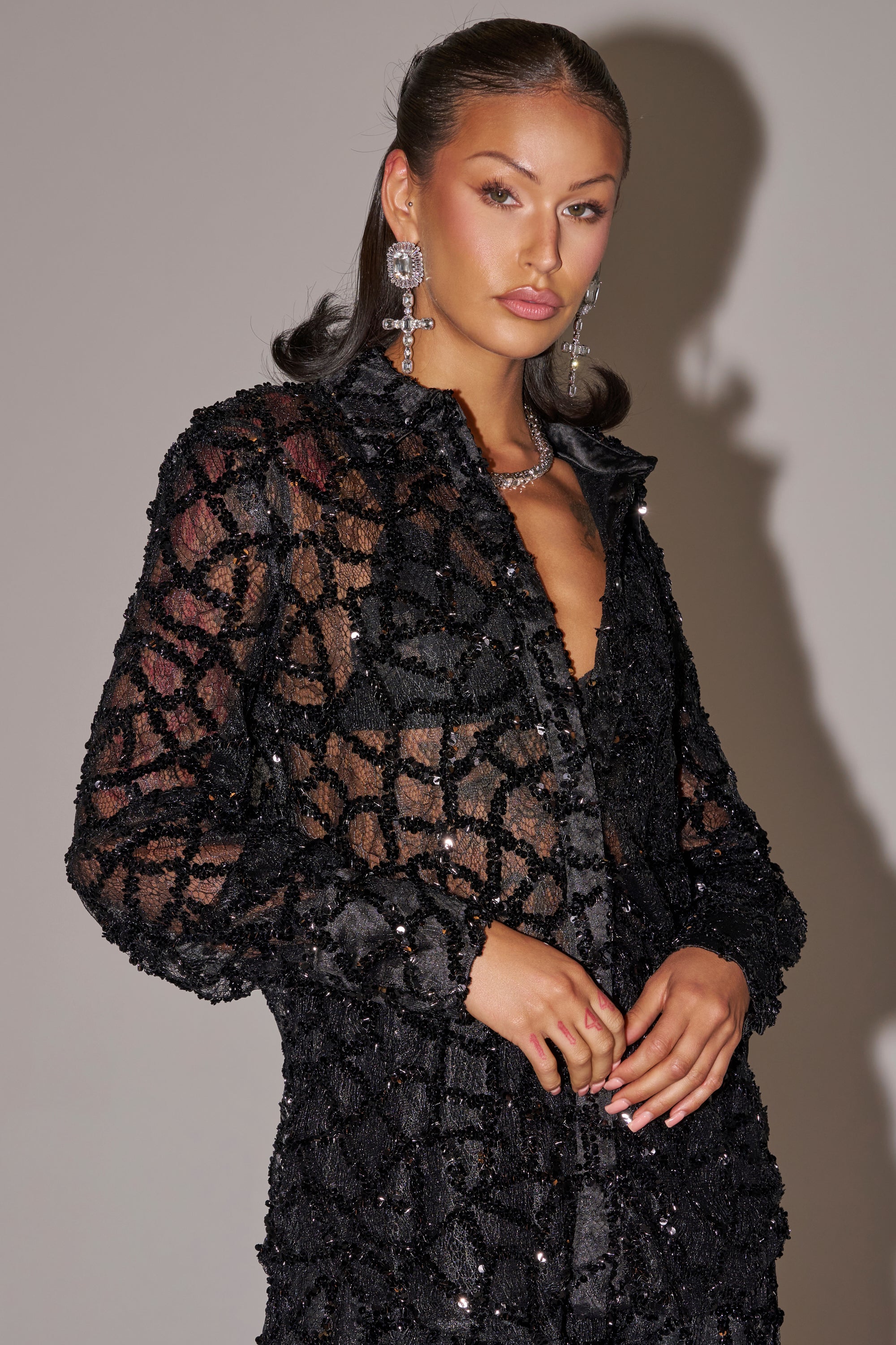 A woman with long brown hair in loose waves wears the ROXY SEQUIN BUTTON DOWN, a sheer black sequin blouse with a geometric pattern, paired with drop earrings and a necklace, standing against a neutral background.