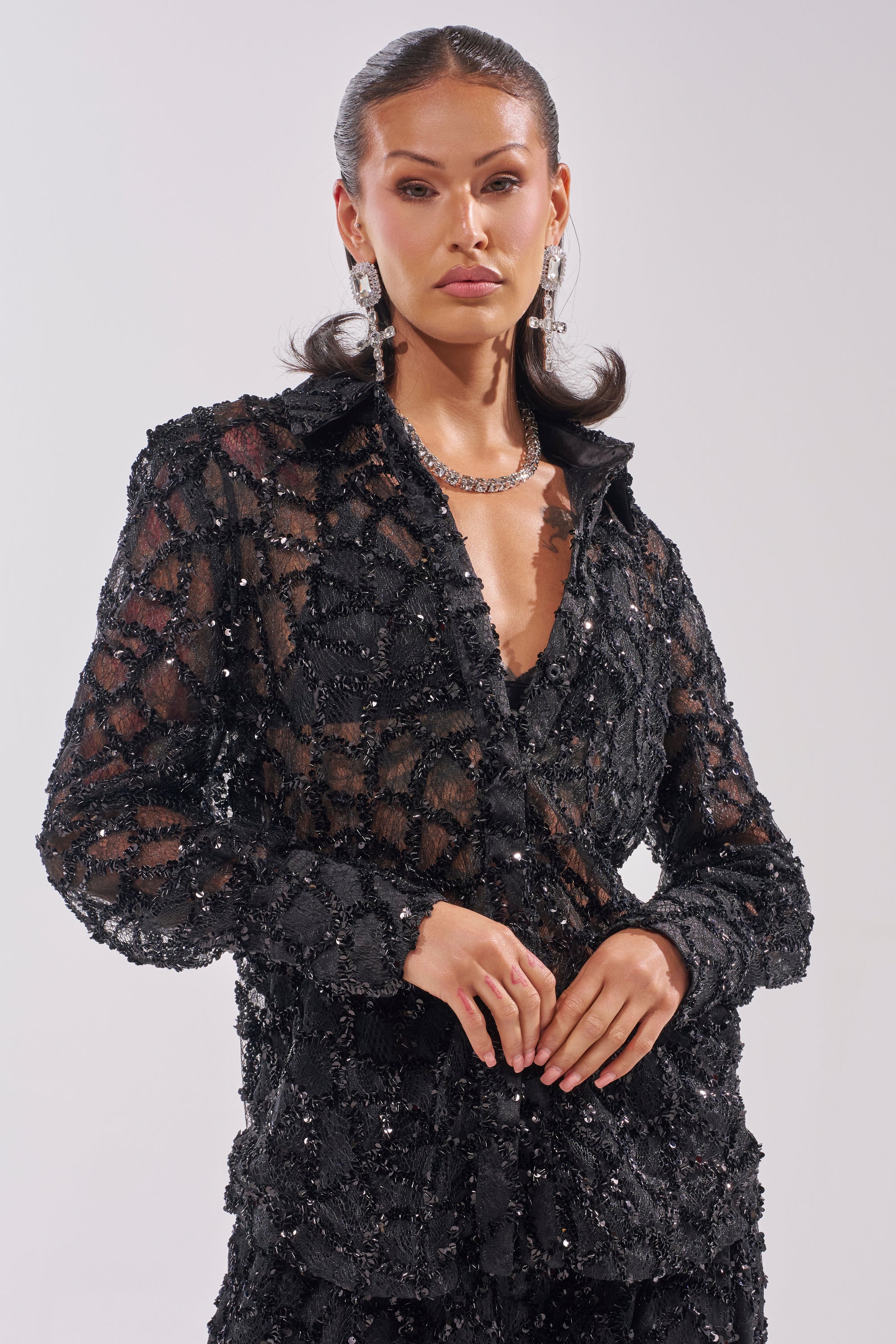 A woman poses against a plain light background wearing the ROXY SEQUIN BUTTON DOWN—a black, sheer blouse with beaded details and a deep neckline—styled with slicked-back hair, statement earrings, and a necklace for an elegant evening look.
