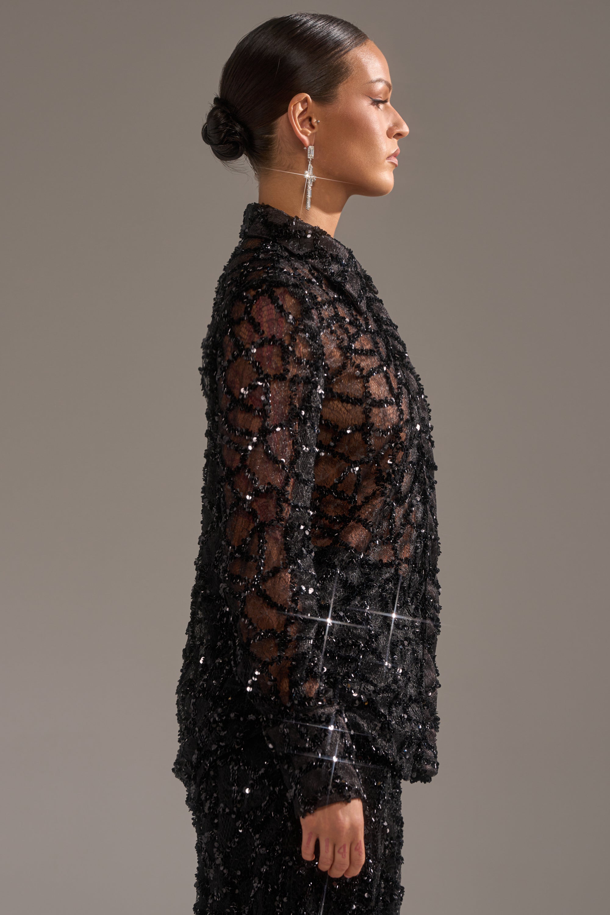 A woman with dark hair in a low bun, wearing the ROXY SEQUIN BUTTON DOWN and long earrings, stands in profile against a plain gray background for an elegant evening look.