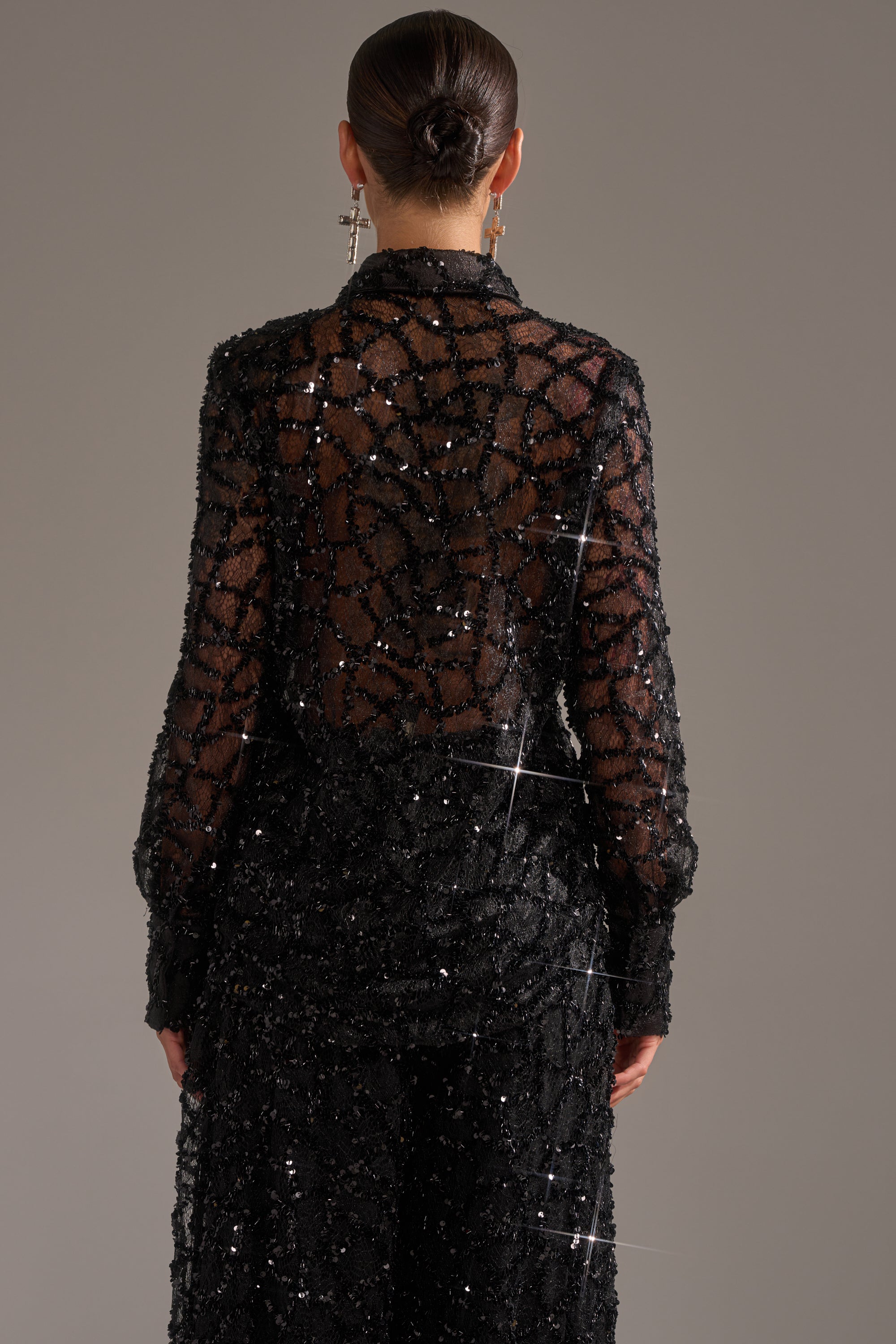 A person with dark hair in a low bun models the ROXY SEQUIN BUTTON DOWN, a sheer blouse adorned with black geometric sequins, shown from the back against a plain gray background for an elegant evening look.