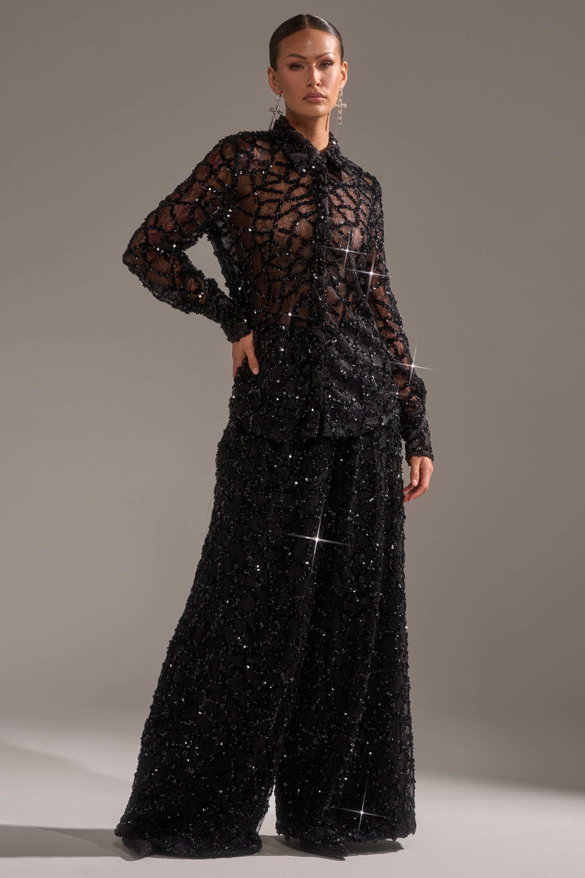 A woman stands confidently in the ROXY SEQUIN BUTTON DOWN blouse and wide-leg black sequin pants, her hair slicked back and accessorized with dangling earrings against a neutral background.