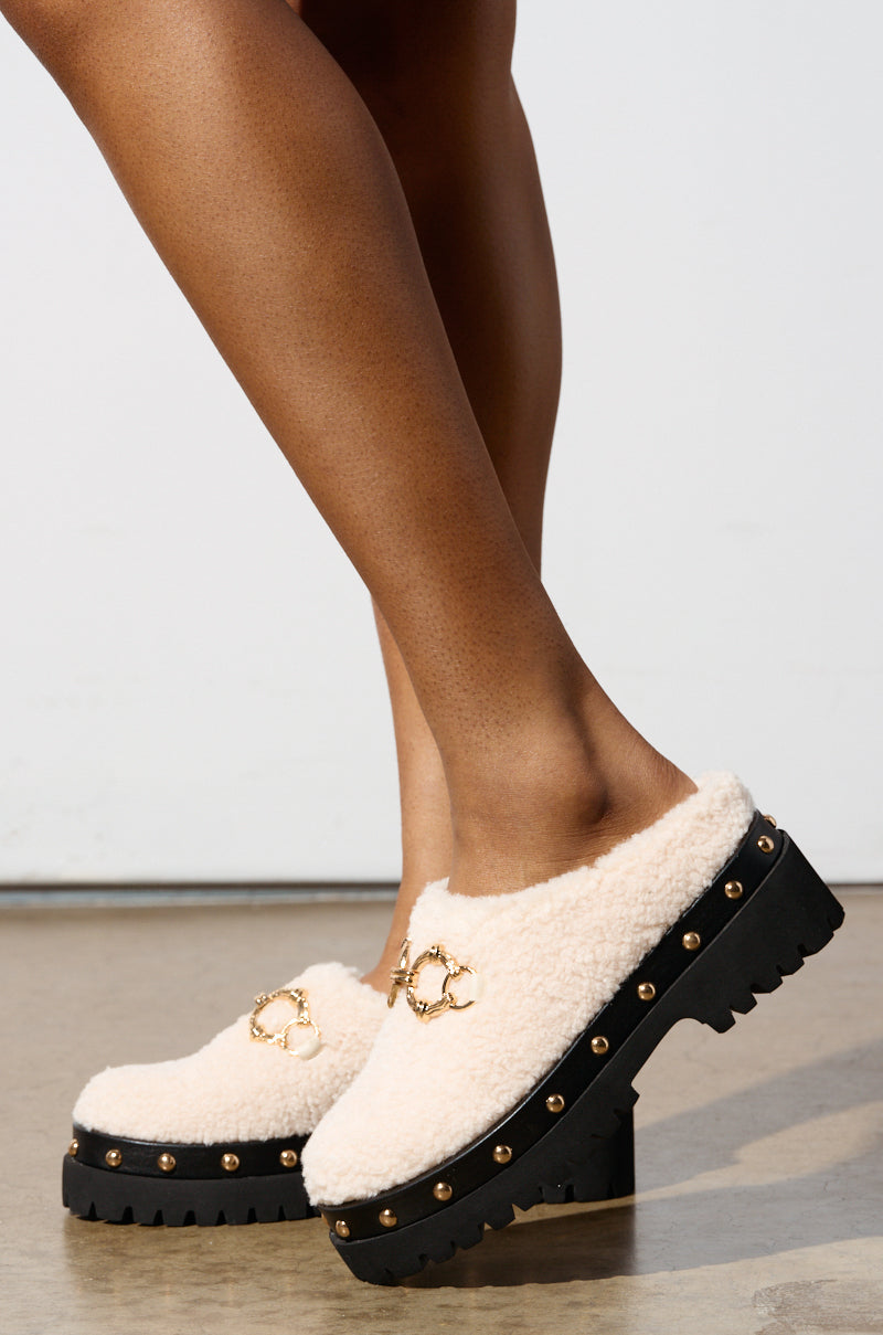 A person stands on a neutral background wearing the AZALEA WANG ROZELE BEIGE CLOGS, showcasing their faux sherpa material, chunky black platform lug soles, and gold chain accents.