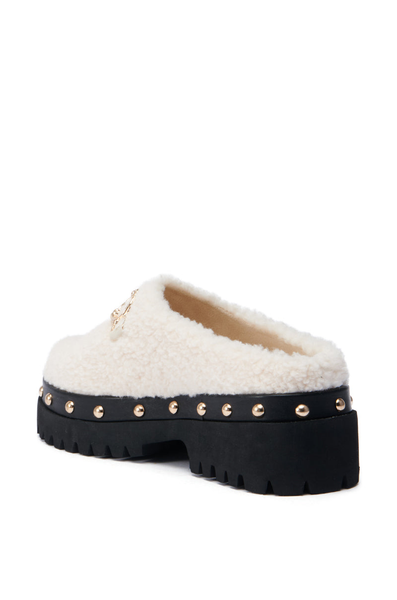 The AZALEA WANG ROZELE BEIGE CLOG is a white faux sherpa slipper with a bold black platform lug sole, accented by gold studs around the base and equipped with a thick tread for enhanced grip.