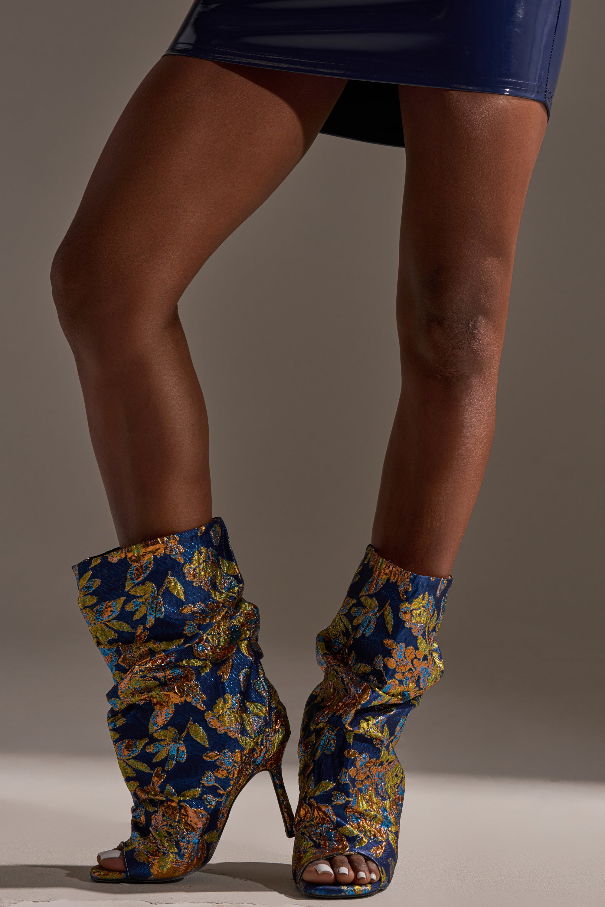 A person wears the AZALEA ROZSI BLUE MULTI BROCADE OPEN TOE STILETTO BOOTIE with a short, shiny dark skirt, standing with one leg crossed in front of the other against a neutral background.