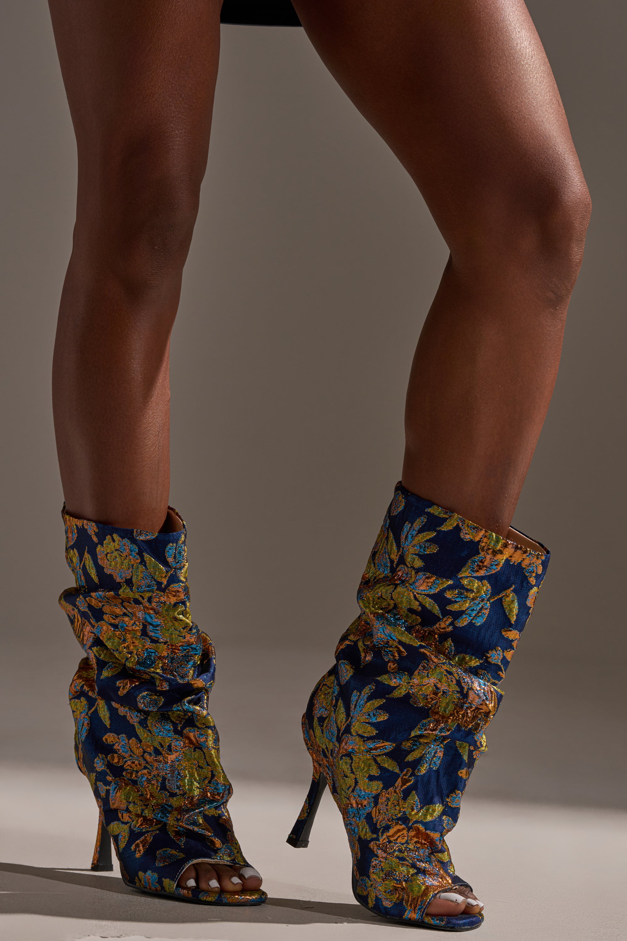 Wearing the AZALEA ROZSI BLUE MULTI BROCADE OPEN TOE STILETTO BOOTIE, featuring a blue and gold floral pattern, a person in a black skirt stands with one leg slightly forward against a neutral background.