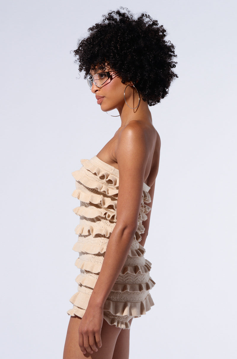 A woman with an afro hairstyle is wearing glasses and the RUFFLES ALL OVER ROMPER, a strapless beige mini dress featuring a bodycon fit and multiple layers of ruffles. She is standing in profile against a plain light background.