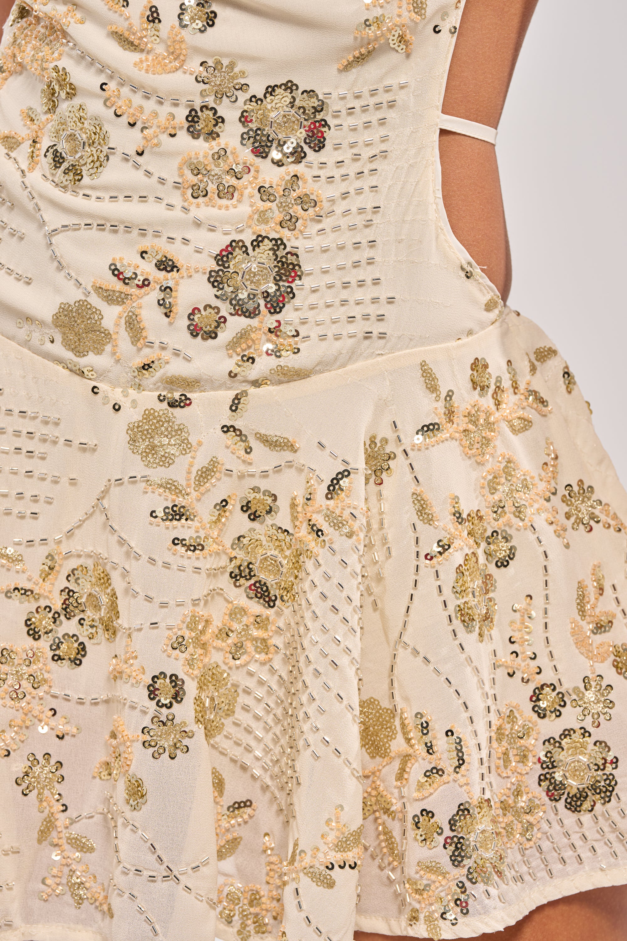 Close-up of the FELICITY EMBELLISHED MINI DRESS, a cream mini dress with gold and silver sequin floral embroidery, featuring a backless design, cut-out waist, and thin white shoulder strap. 7