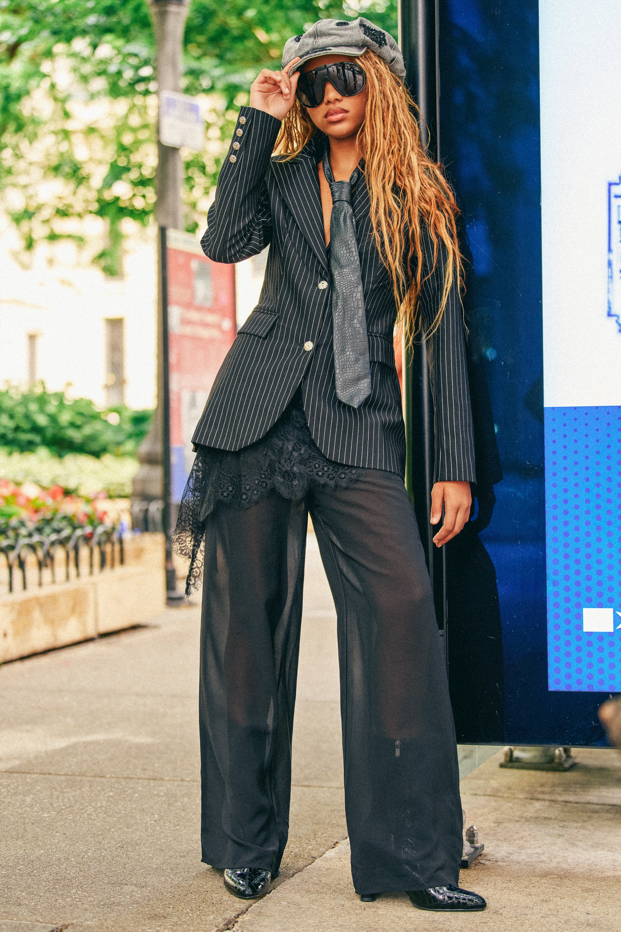 A stylish person poses outdoors in the DRIFT AWAY PALAZZO PANT WITH LACE SKIRT OVERLAY, paired with a pinstripe blazer and tie. Sheer details, sunglasses, and a gray newsboy cap complete the look against a backdrop of trees and urban scenery.