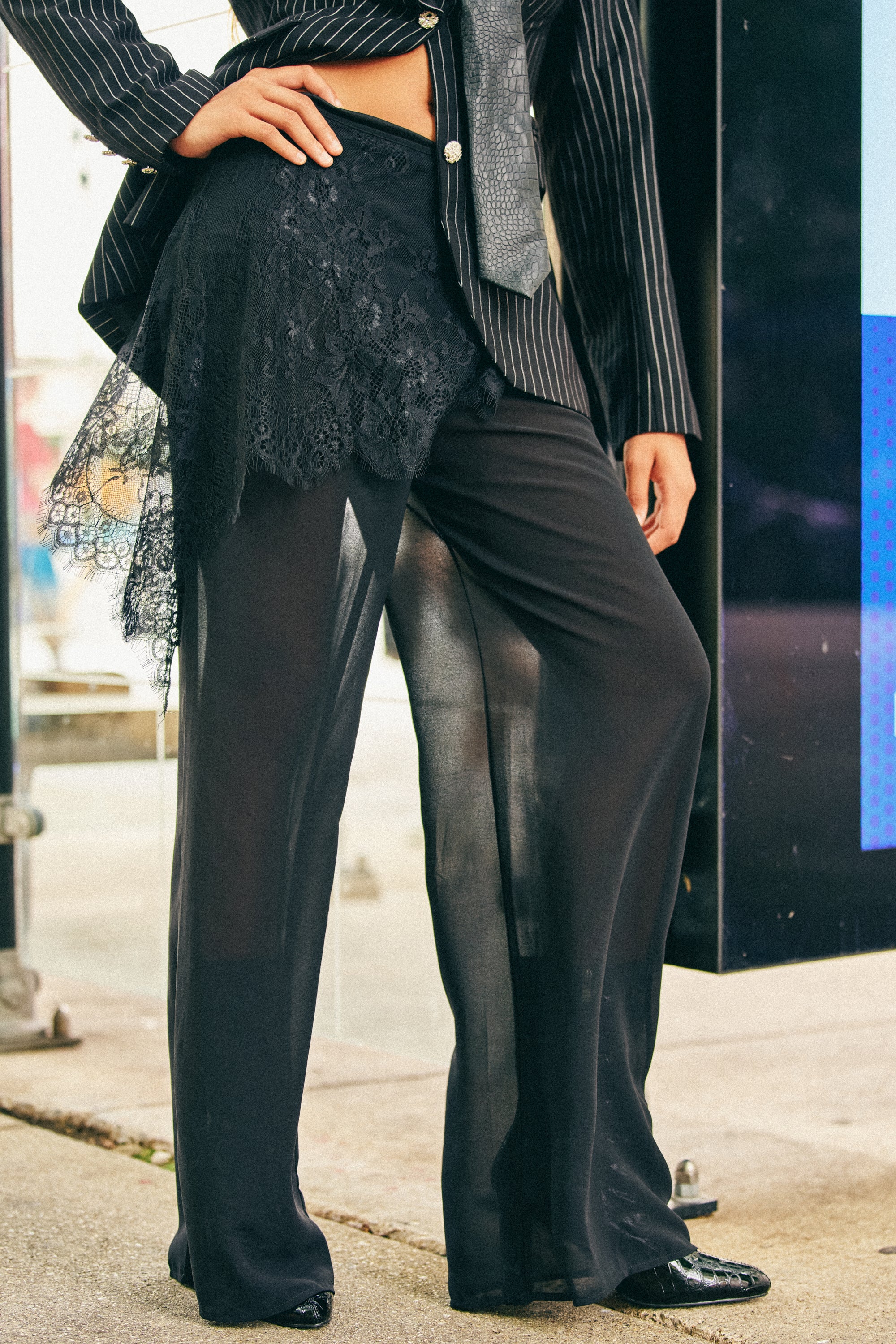 Wearing the DRIFT AWAY PALAZZO PANT WITH LACE SKIRT OVERLAY and a pinstripe blazer, this person stands outdoors on a sidewalk. The outfit features a striking wide-leg silhouette with sheer fabric and lace detailing, showcasing a blend of modern textures and layers.