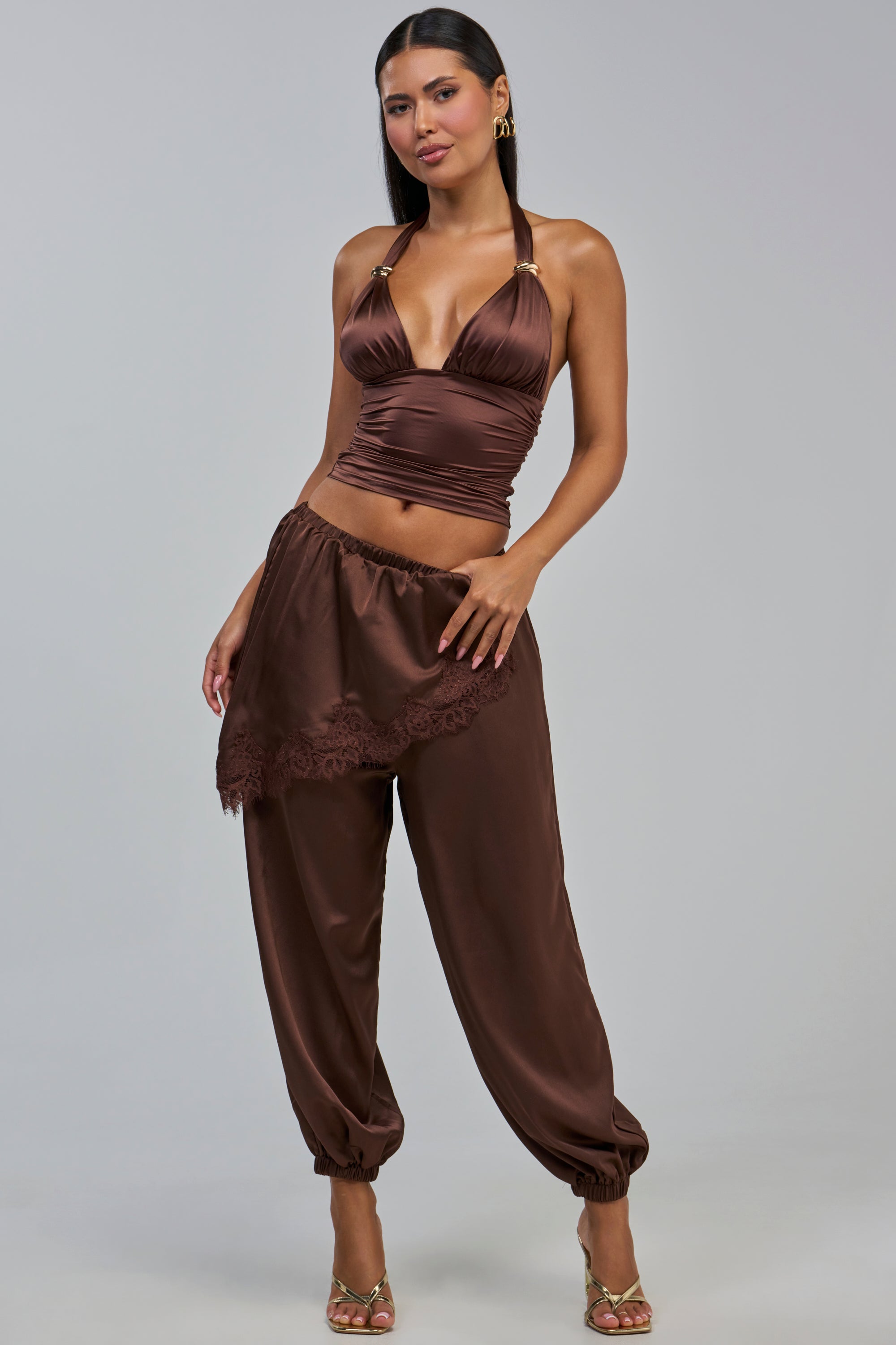 A woman poses in the PARTY IN THE FRONT BALLOON PANT WITH SKIRT OVERLAY, featuring sleeveless, deep v-neck styling and a rich brown satin finish. The matching balloon pants are accented with an asymmetrical, lace-trimmed skirt overlay. Styled with gold sandals and statement earrings, the look radiates boho-chic elegance against a gray background.