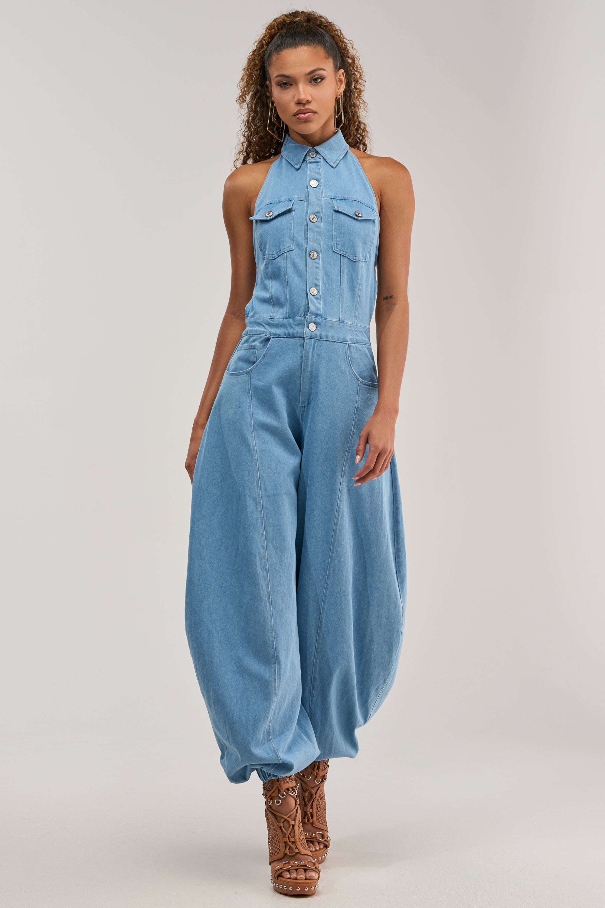 A woman wears the JETSETTER JUMPSUIT IN LIGHT BLUE DENIM, a sleeveless, wide-leg denim jumpsuit, paired with brown strappy heels as she stands against a plain background.