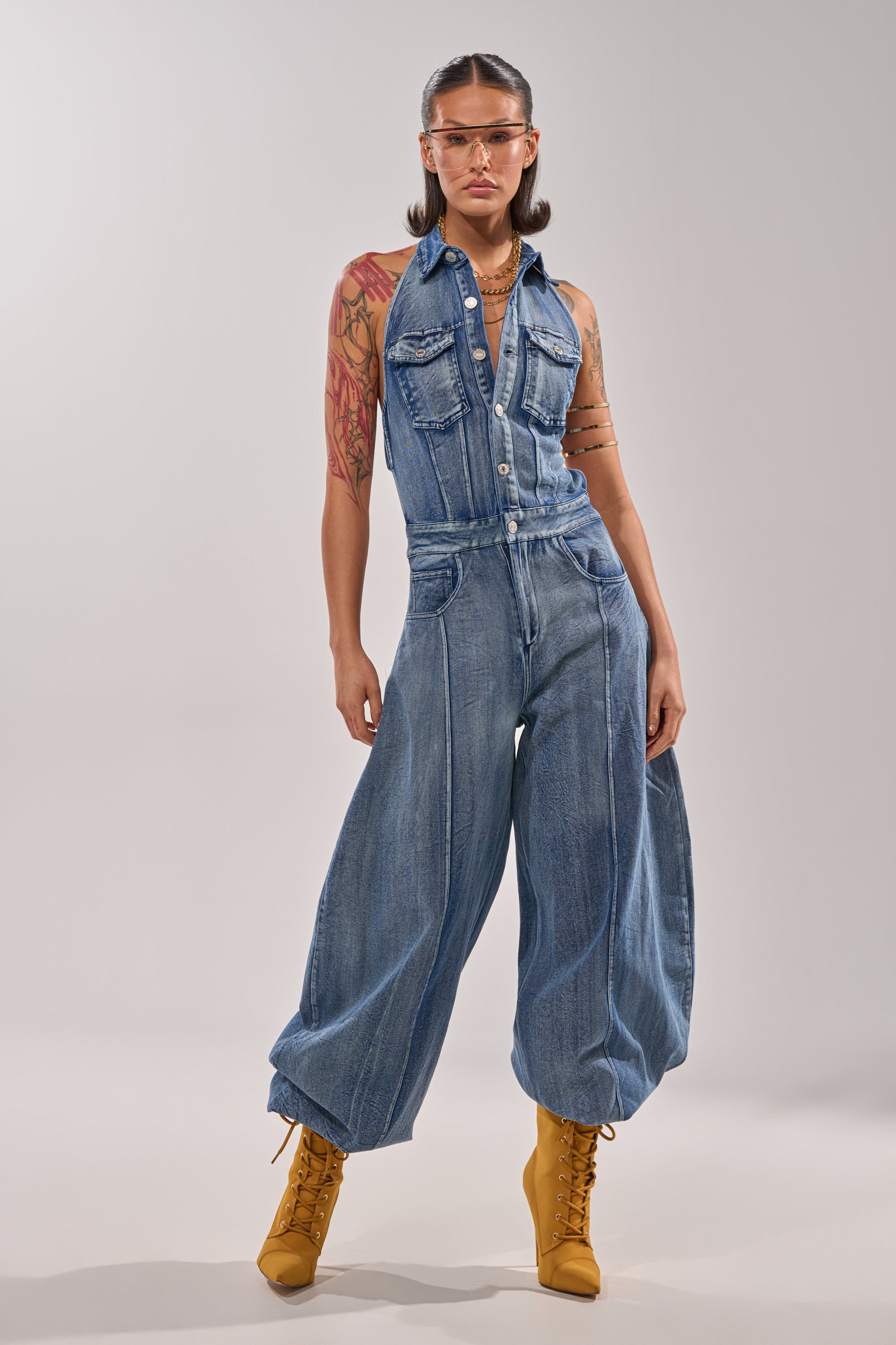 A person wearing the JETSETTER JUMPSUIT IN MEDIUM BLUE DENIM, gold lace-up boots, clear glasses, gold jewelry, and showing arm tattoos stands facing forward against a plain light background. 0