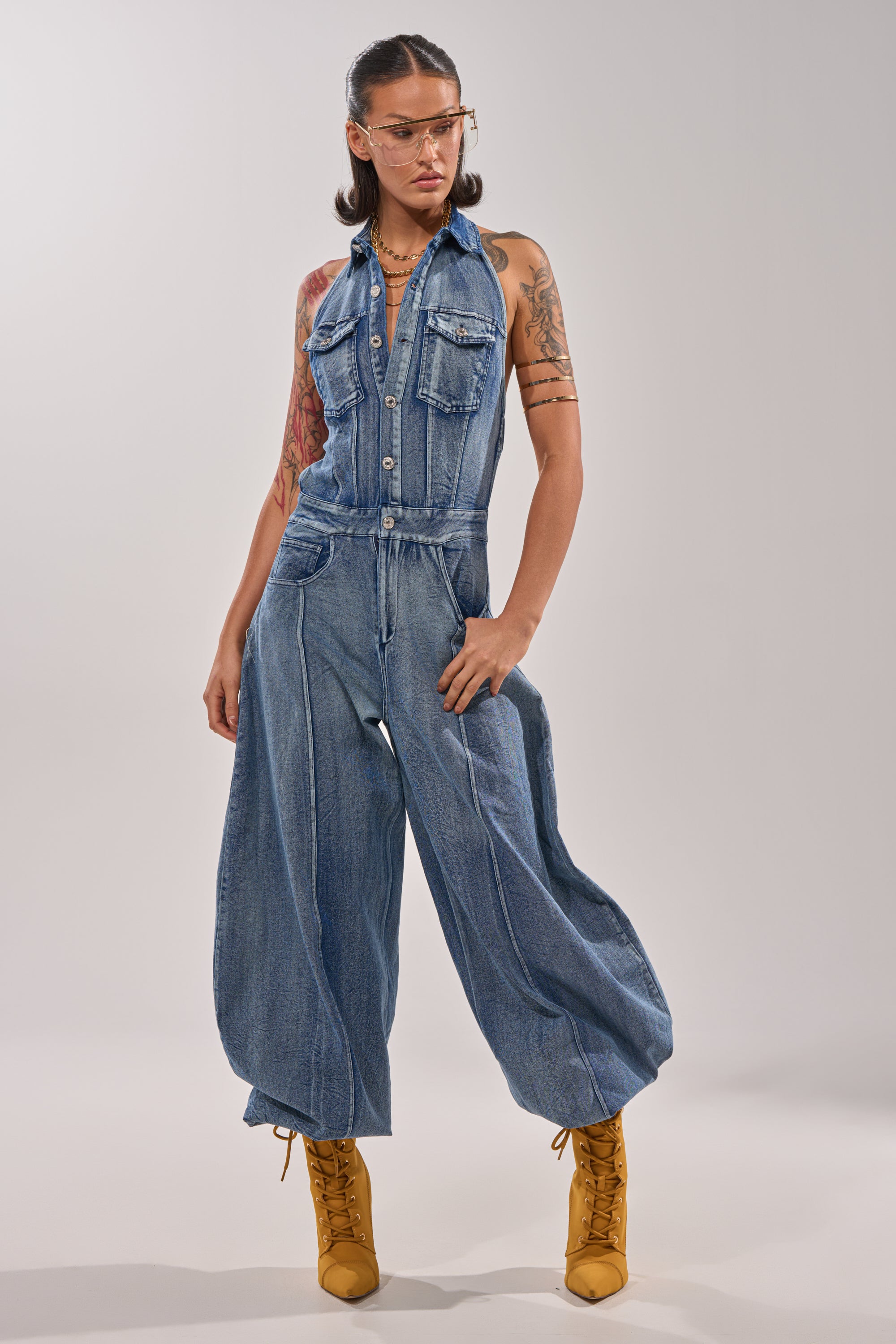 A person stands confidently in a light studio wearing the JETSETTER JUMPSUIT IN MEDIUM BLUE DENIM with wide legs, yellow lace-up boots, clear glasses, layered necklaces, and visible arm tattoos. 1
