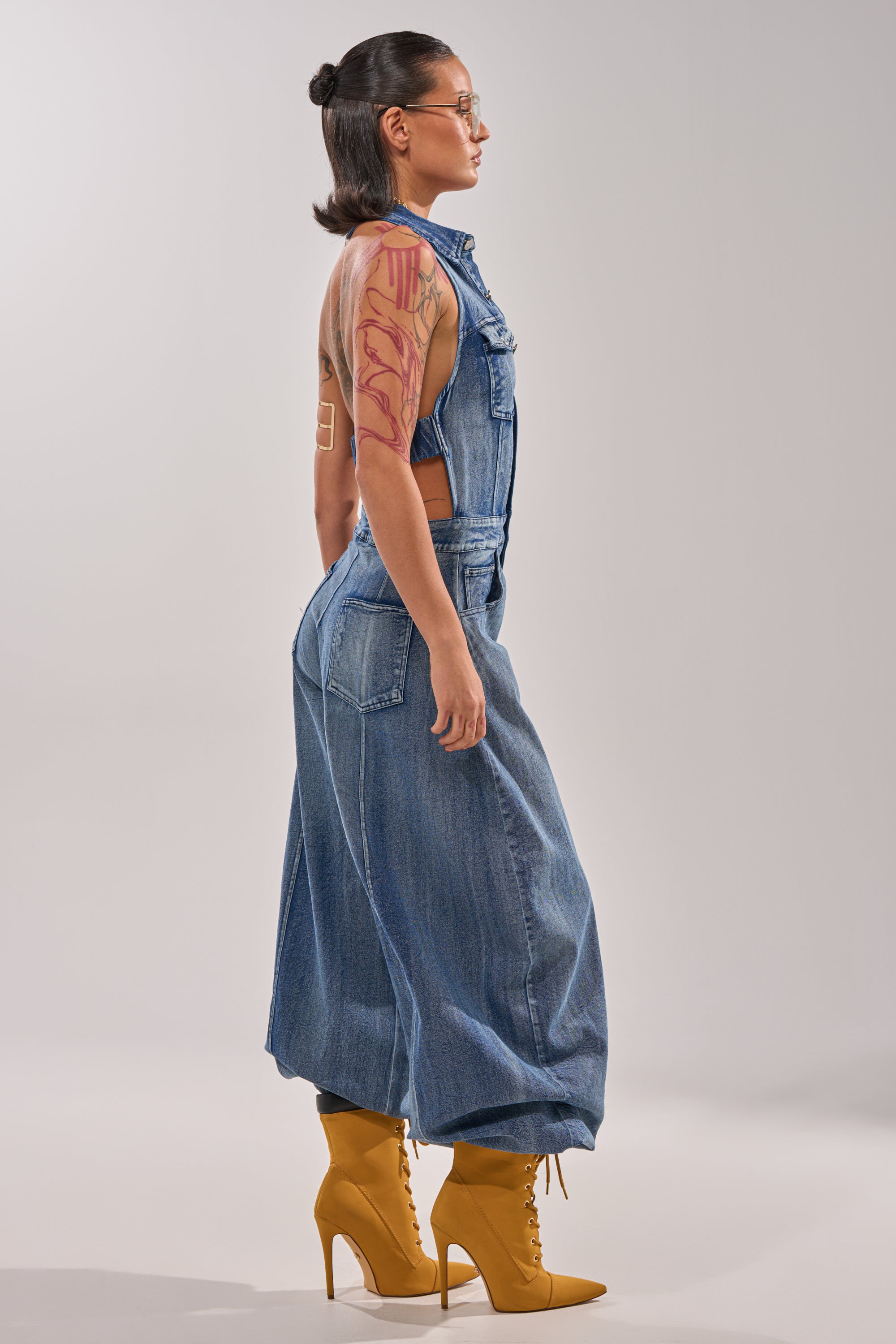 A person with long dark hair in a bun, glasses, and visible arm tattoos poses in profile against a plain background, wearing the JETSETTER JUMPSUIT IN MEDIUM BLUE DENIM paired with yellow high-heeled lace-up boots. 2
