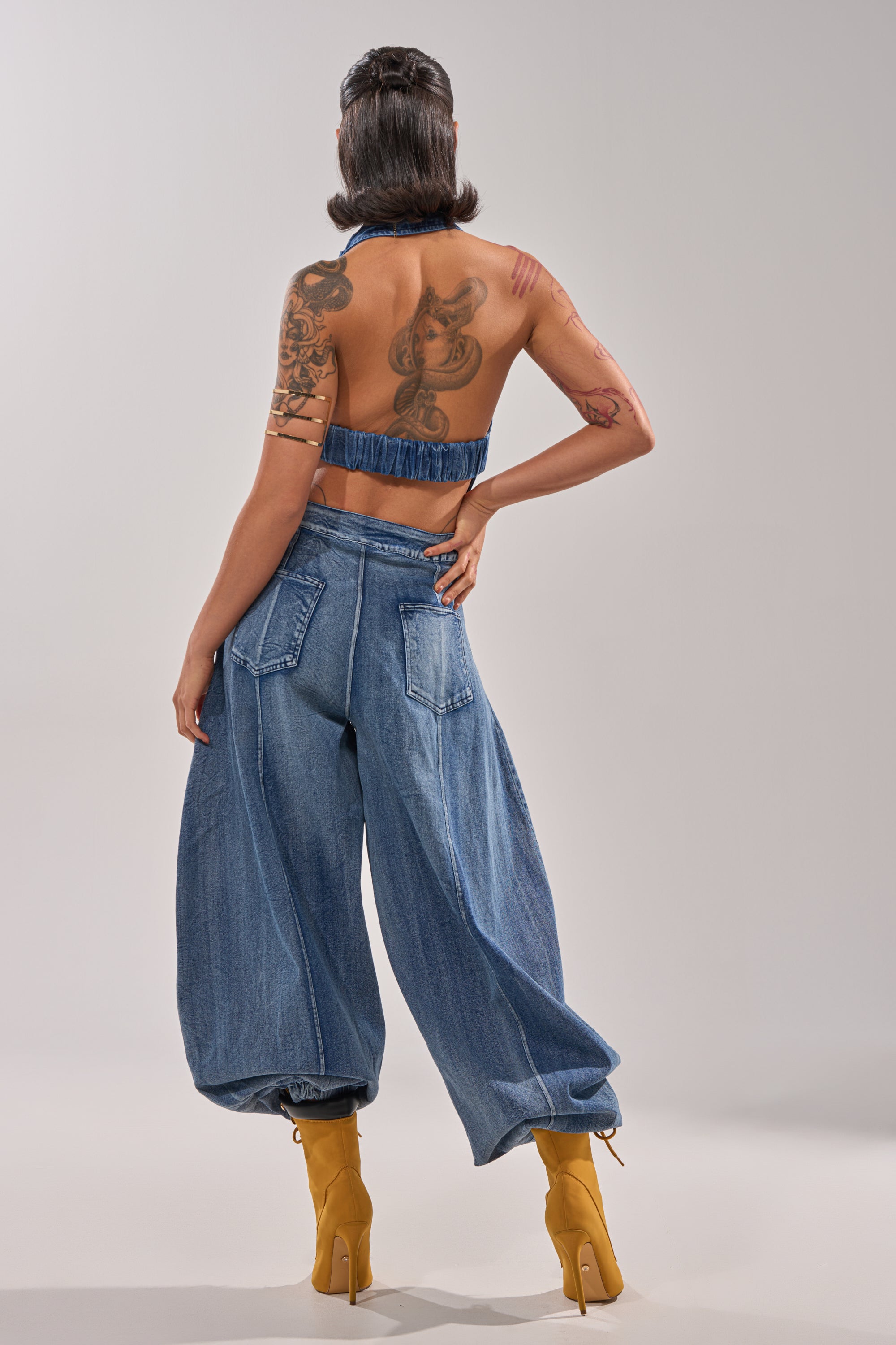 A person stands facing away, wearing the JETSETTER JUMPSUIT IN MEDIUM BLUE DENIM with relaxed utility vibes and tan platform boots. Their arm and back tattoos are visible, and a short haircut completes the look against a plain background. 3