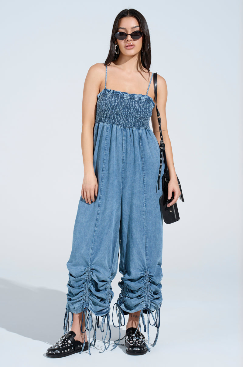 The woman confidently poses against a light blue background, rocking the JUMPIN AROUND DENIM JUMPSUIT with ruched leg details, black studded shoes, and a chic black handbag—all while wearing sunglasses.