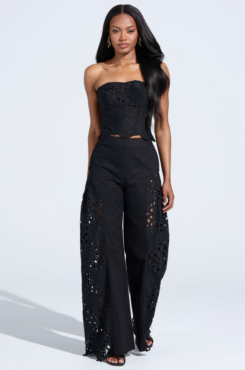 A woman poses against a plain background in the stylish YOU WILL SEE WOVEN TROUSER, a strapless black lace jumpsuit with eyelet wide-leg pants. Her long, dark hair is worn down, complemented by drop earrings and open-toed shoes.