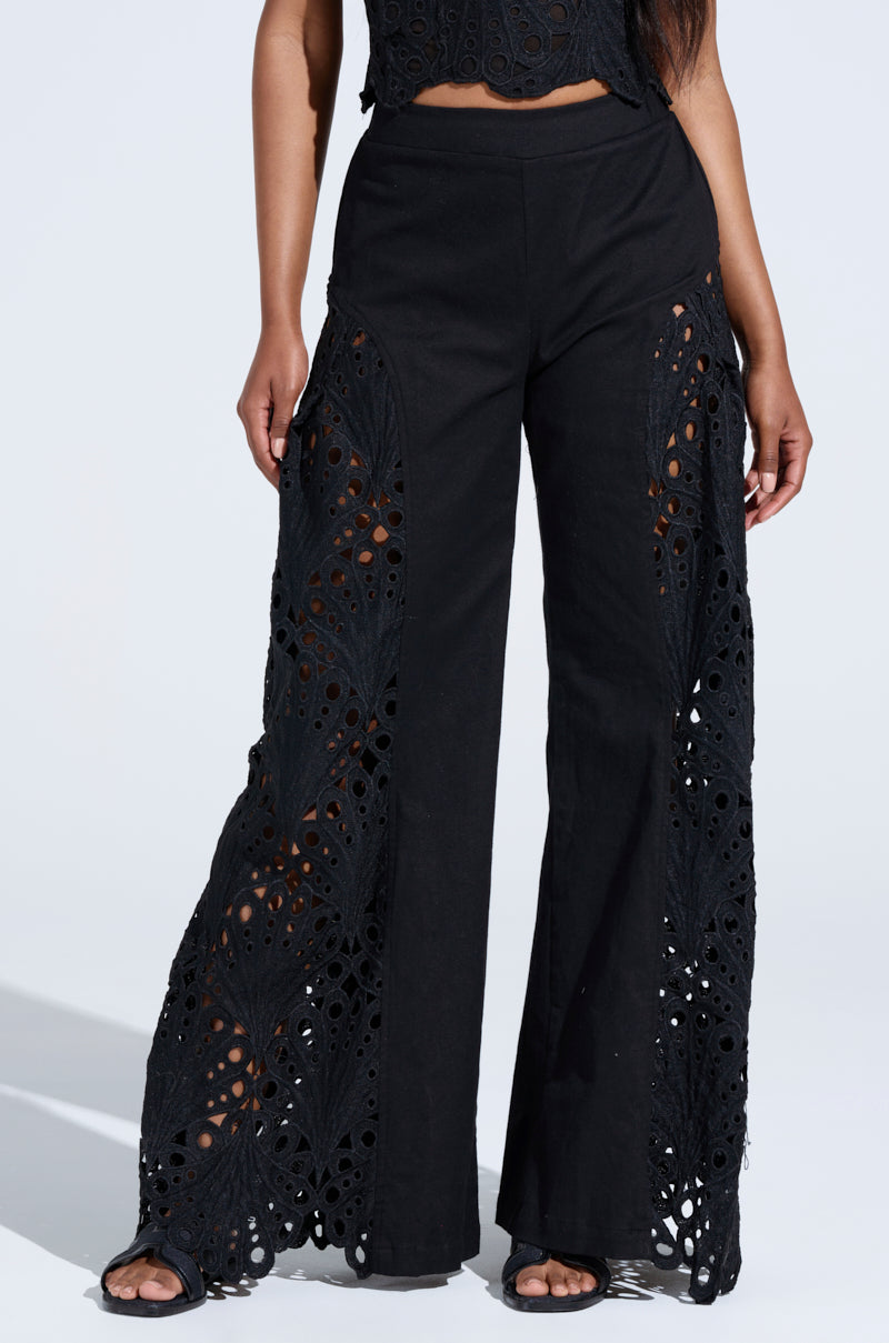 A person is wearing YOU WILL SEE WOVEN TROUSER in black, featuring wide legs and intricate floral lace designs down the sides, paired with black sandals. The trousers are high-waisted against a plain, light-colored background.