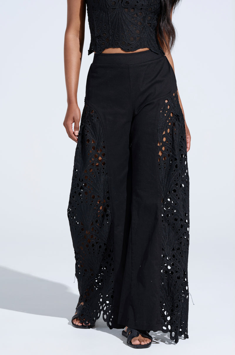 A person is wearing a black lace ensemble with a sleeveless top and YOU WILL SEE WOVEN TROUSER, featuring intricate cut-outs and side splits accented by lace patterns, all poised against a light background.