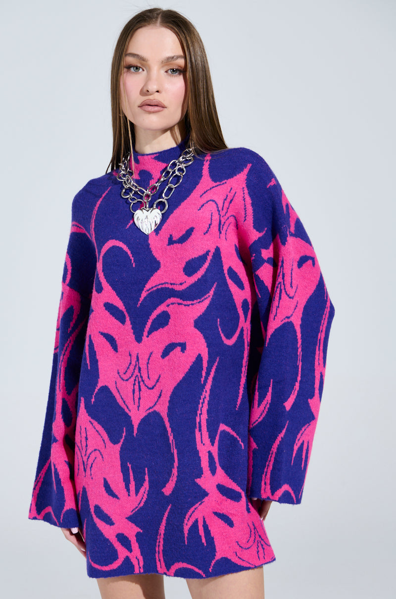 A woman wears the MUSHU Graphic Sweater Dress in blue and pink, featuring abstract patterns. She accessorizes with a large silver chain necklace with a heart pendant, while her straight hair cascades past her shoulders against a plain light gray backdrop.