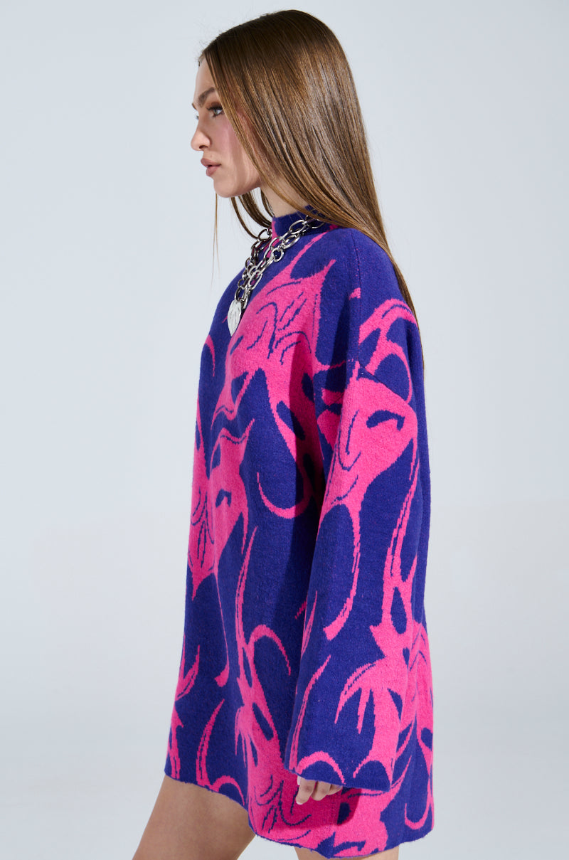 A woman with long brown hair in profile wears the MUSHU GRAPHIC SWEATER DRESS IN BLUE AND PINK. She accessorizes with a chunky silver necklace, standing gracefully against a plain background.