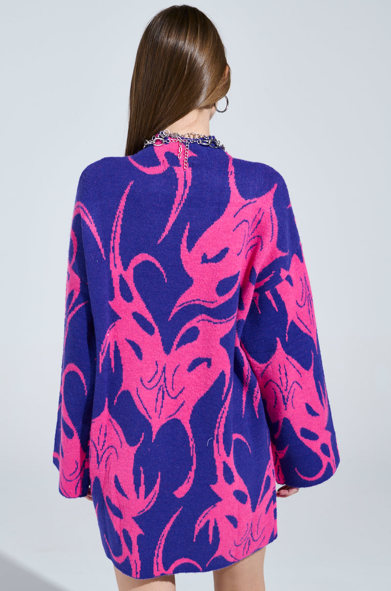 A woman is wearing the MUSHU Graphic Sweater Dress in blue and pink, featuring an oversized silhouette with a tribal pattern. She stands with her back to the camera, showcasing its loose fit and long sleeves.