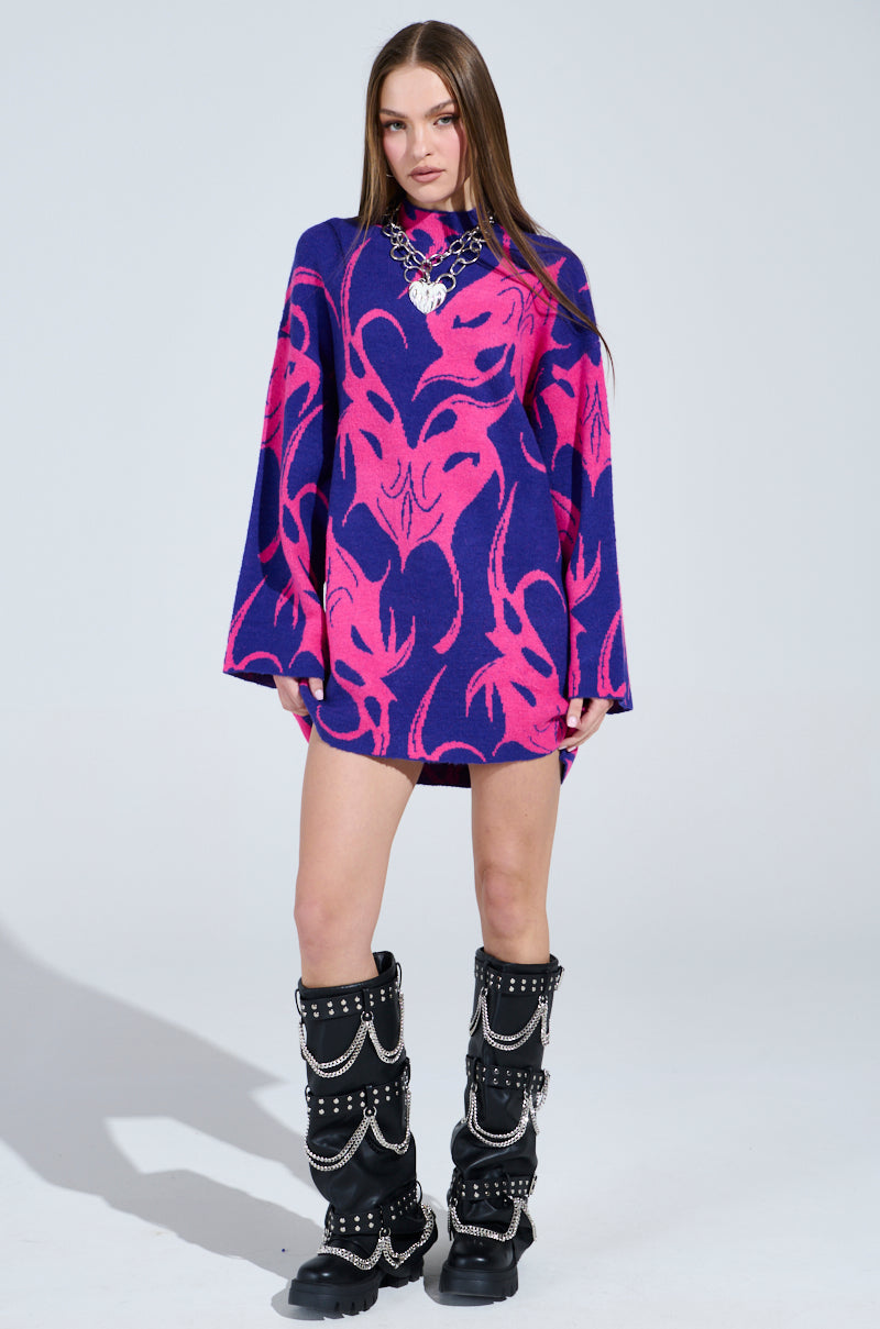 A person confidently wears the MUSHU GRAPHIC SWEATER DRESS IN BLUE AND PINK, featuring a vibrant abstract pattern. Their long hair flows over the oversized silhouette, while chunky black boots with chains complete the look against a plain, light-colored background.