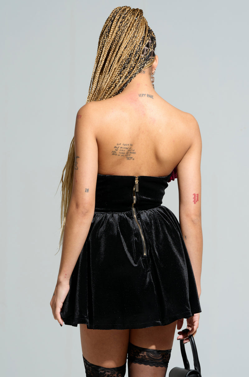 Seen from the back, a person with long braided hair wears the LOVER GIRL ERA VELVET FLOWER EMBELLISHED MINI DRESS. The outfit features a black strapless design with a corset-inspired bodice and zipper. Tattoos adorn their upper back and arms, and they carry a small handbag. Black lace stockings complete their look.