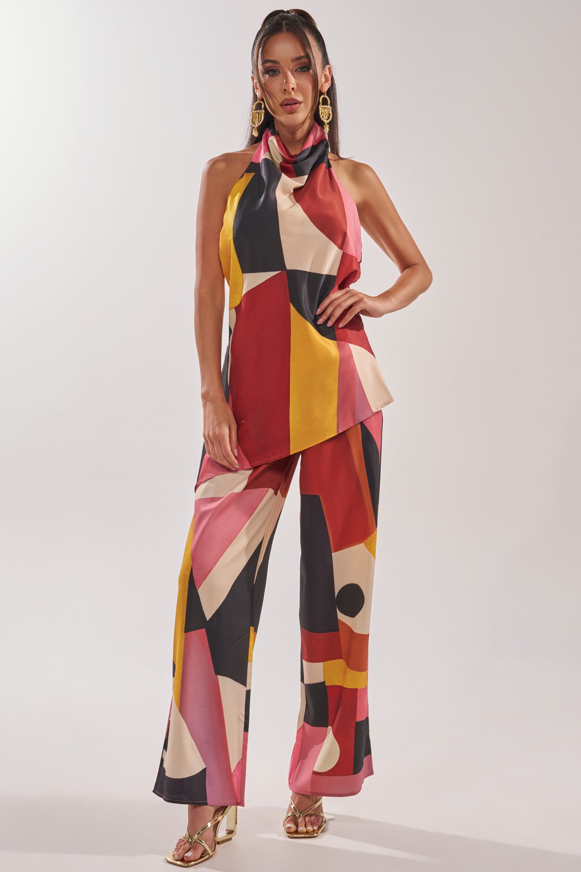 A woman models the MIXED EMOTIONS TROUSER—wide-leg pants with a bold red, yellow, black, and white geometric pattern—paired with a sleeveless halter top, gold earrings, and heels against a plain white background. 0