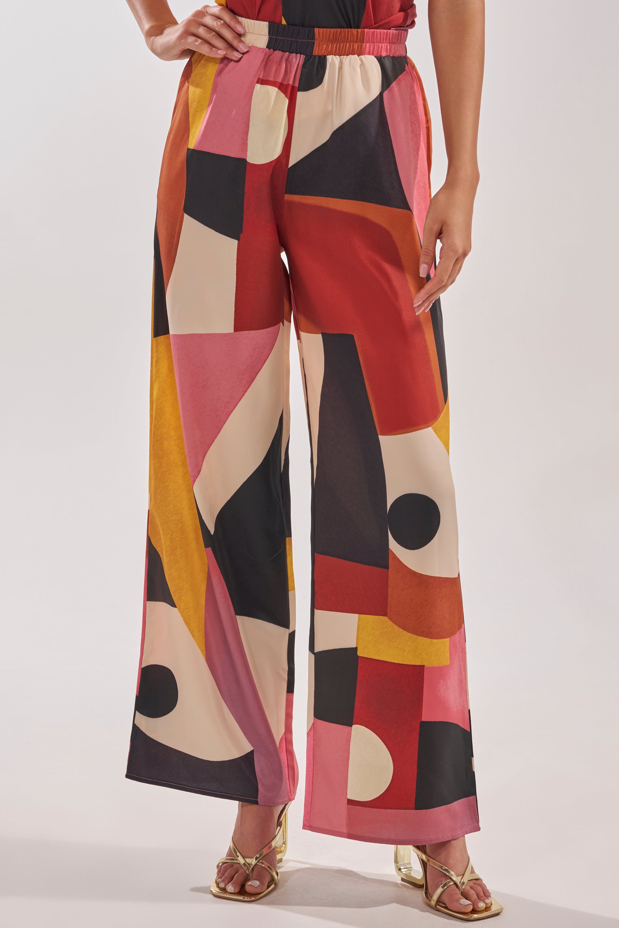 A person styled in the MIXED EMOTIONS TROUSER—statement geometric wide-leg pants in red, pink, yellow, black, and cream—paired with gold open-toe heels. 1
