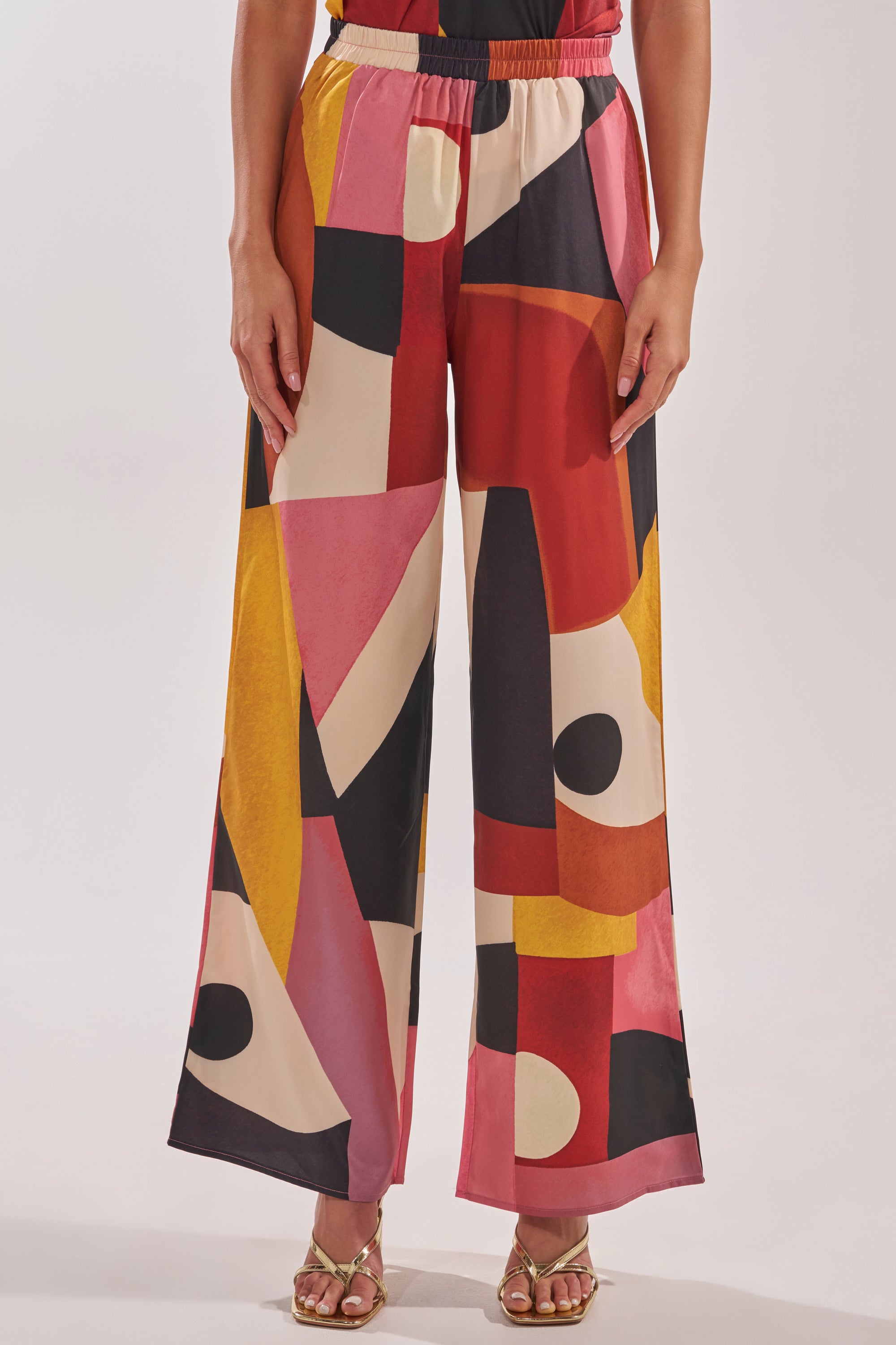 A person wears the MIXED EMOTIONS TROUSER—geometric wide-leg vacation pants with a bold red, yellow, pink, black, and cream pattern—while standing against a plain background. 2