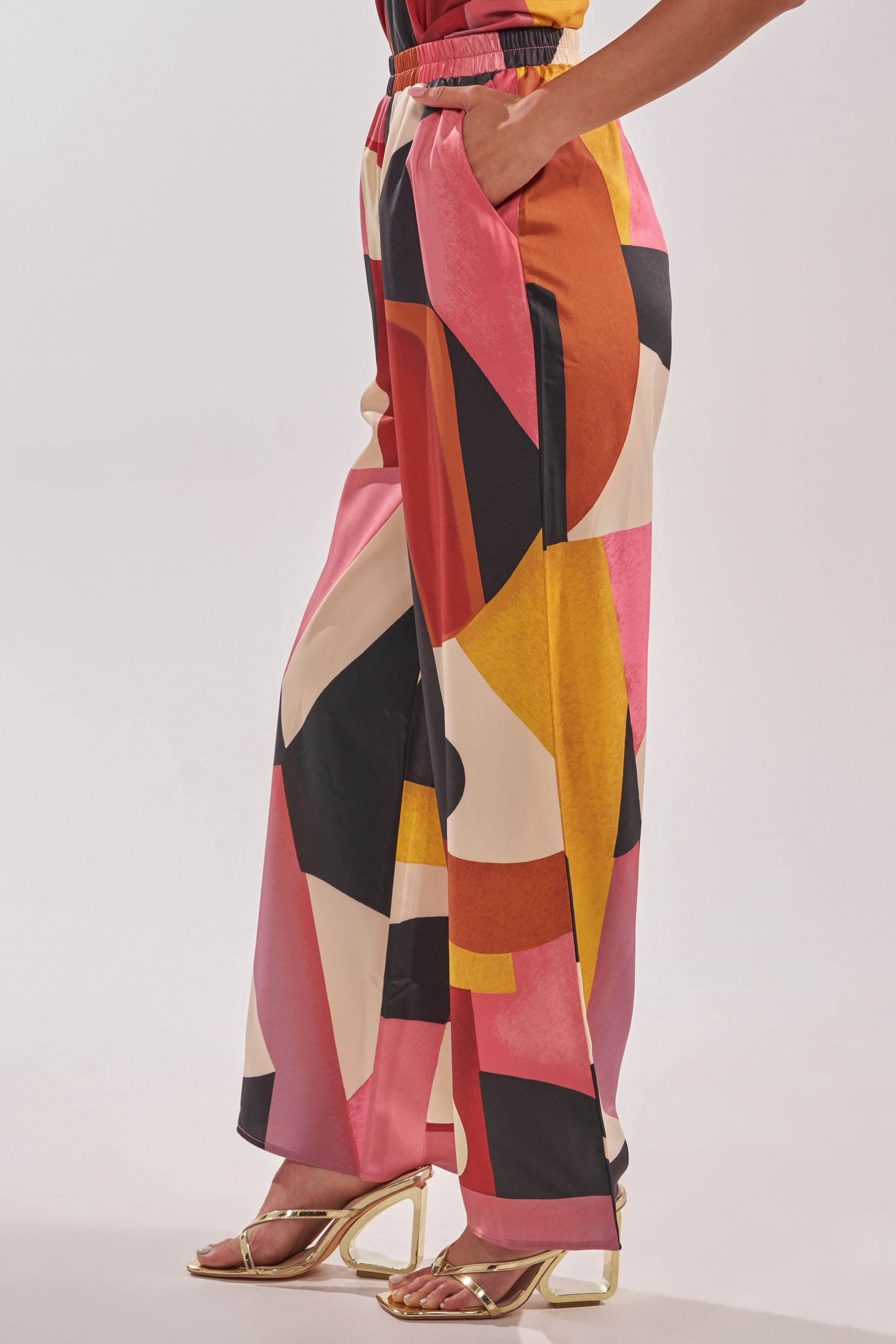Wearing the MIXED EMOTIONS TROUSER, which features a bold geometric pattern in pink, red, yellow, black, and white, paired with gold strappy high heels against a plain background. 3