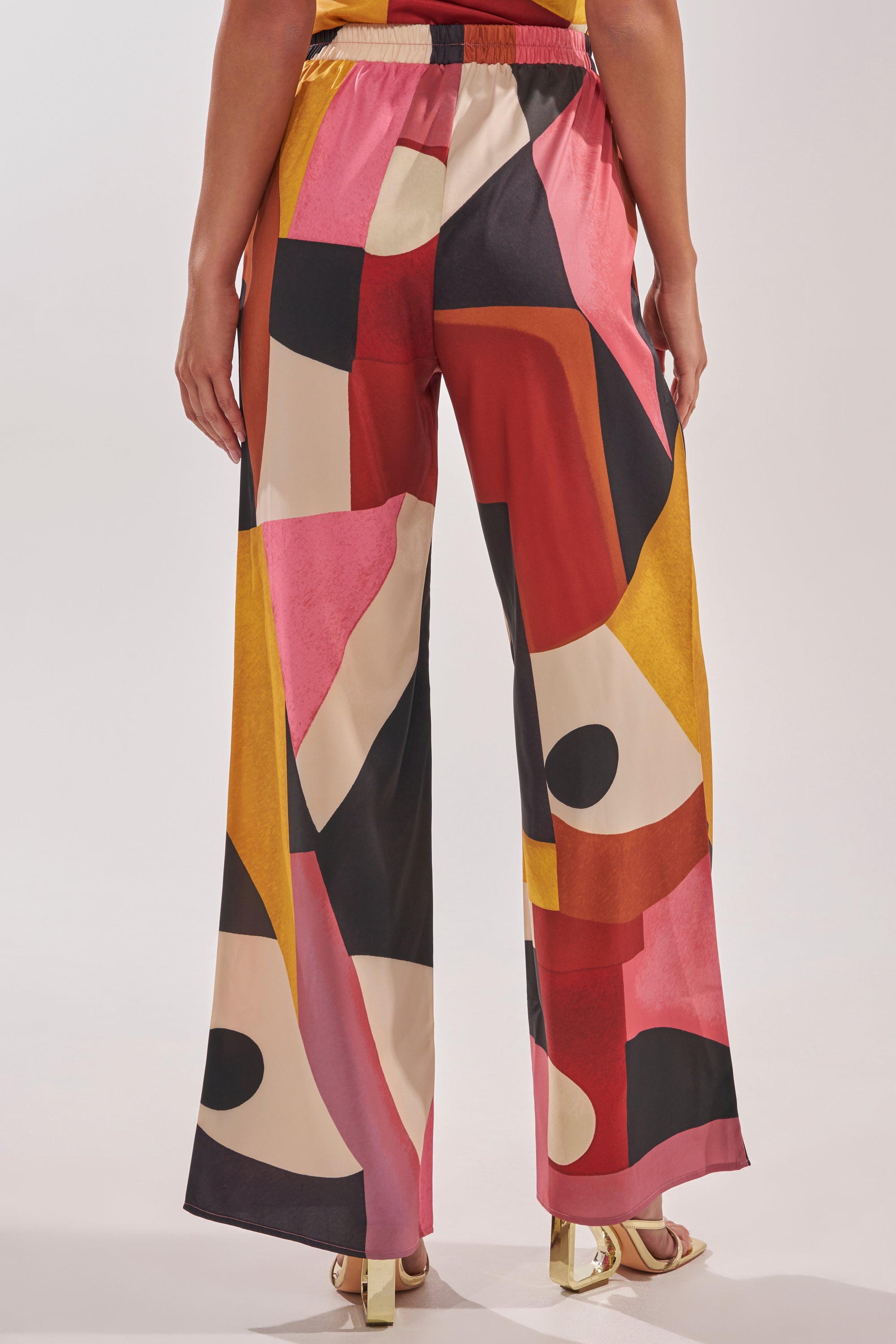A person wearing the MIXED EMOTIONS TROUSER, featuring a bold geometric pattern in red, pink, yellow, black, and cream, stands with their back to the camera. 4