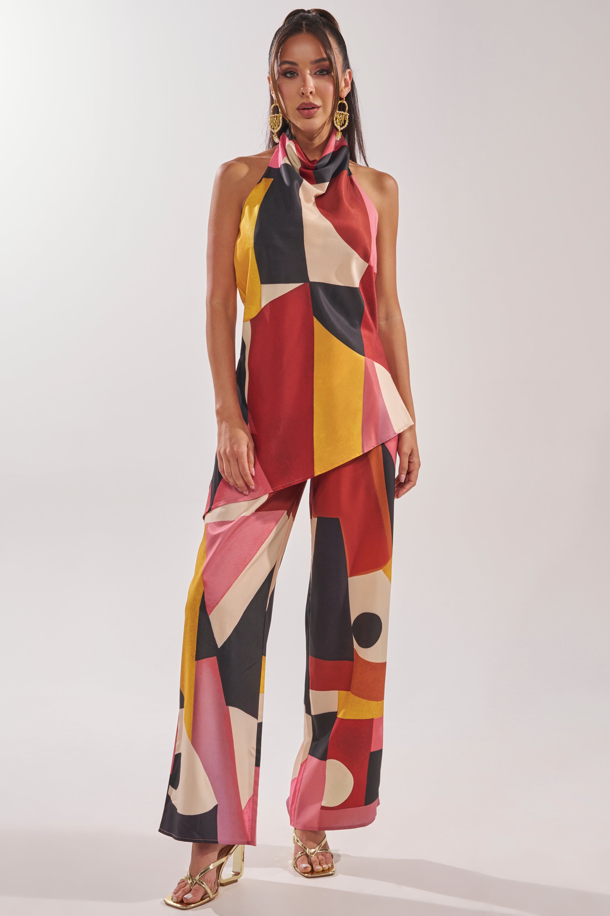 A woman poses in the MIXED EMOTIONS TROUSER paired with a colorful halter top, statement earrings, and gold heeled sandals against a plain light background. 5